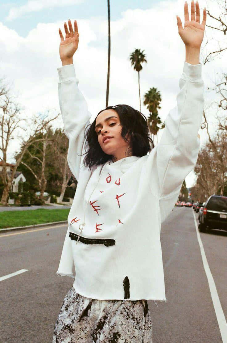 kehlani 桌布 壁紙 — 一位身穿帶有紅色塗鴉的白色連帽衫的年輕女子站在街上，在棕櫚樹和陰天之下舉起雙手。