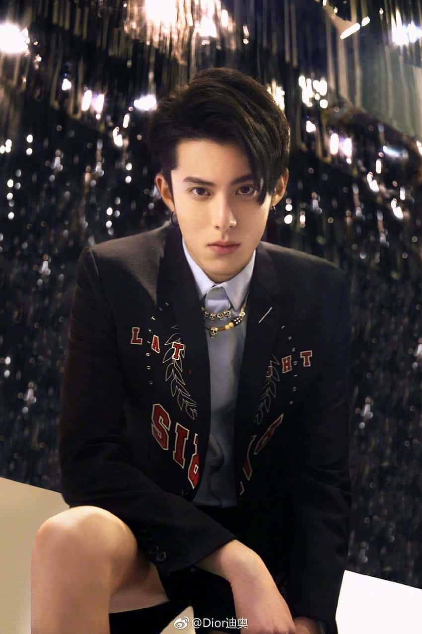 Dylan Wang 桌布 壁紙 — 一位身穿帶有紅色字樣的黑色西裝外套的年輕男子，自信地坐在閃閃發光的黑色窗簾背景前。