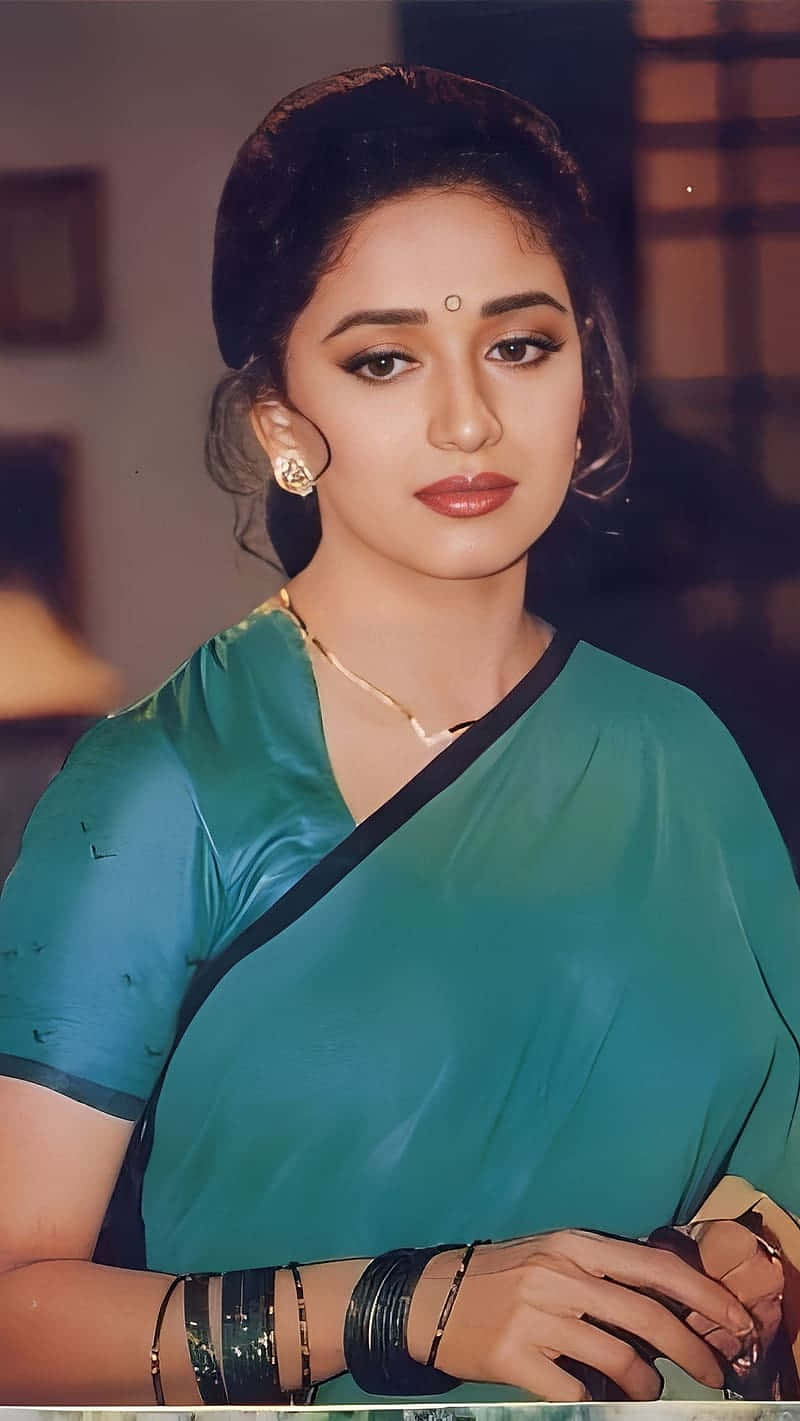  壁紙 — 一位身穿帶黑色飾邊和金色珠寶的藍綠色紗麗的女性，在溫暖的室內環境中溫柔地凝視著相機。