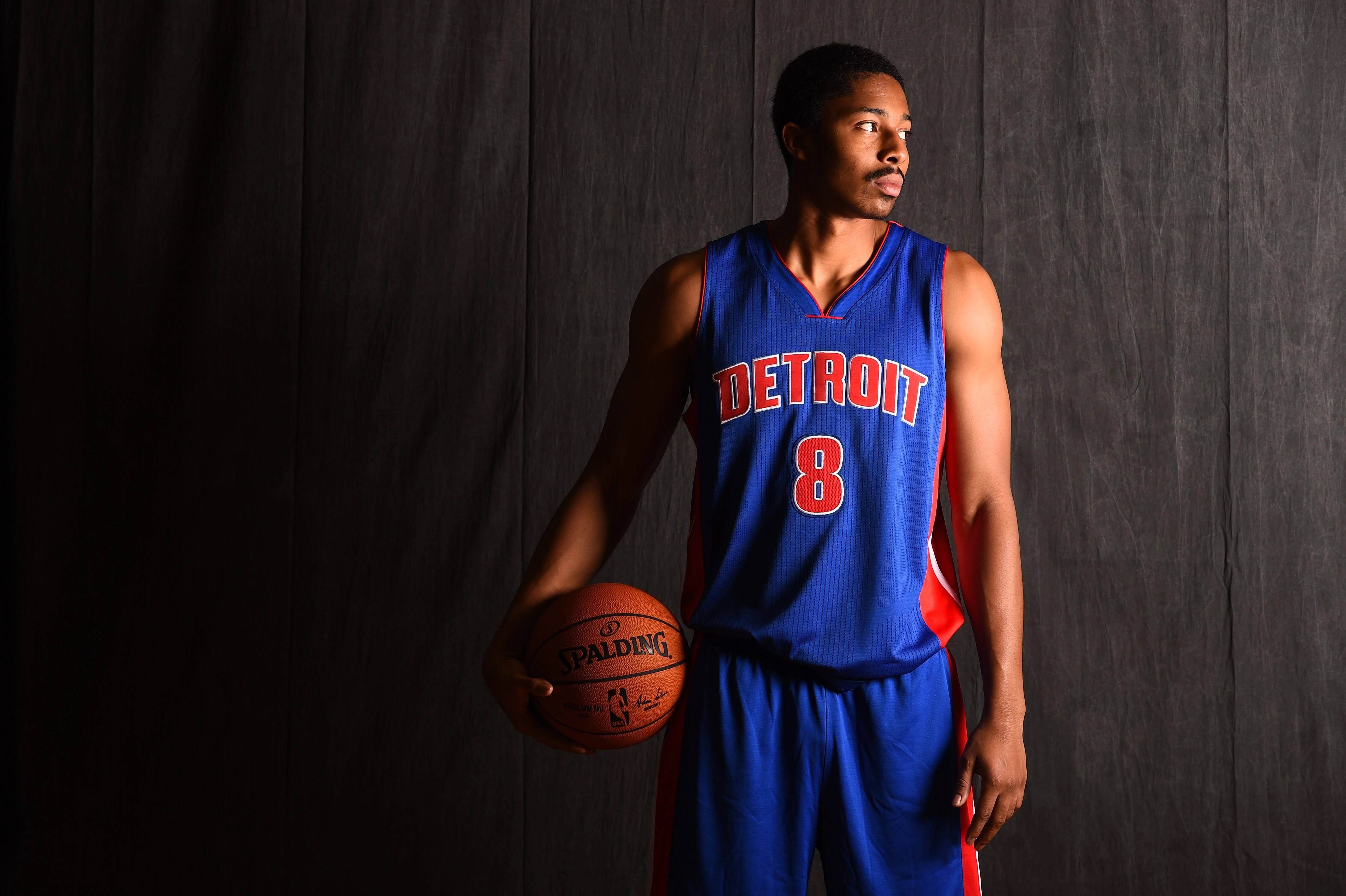 spencer dinwiddie 桌布 壁紙 — 一位身穿底特律活塞隊球衣的籃球運動員，在深色木質背景前拿著一個Spalding籃球。