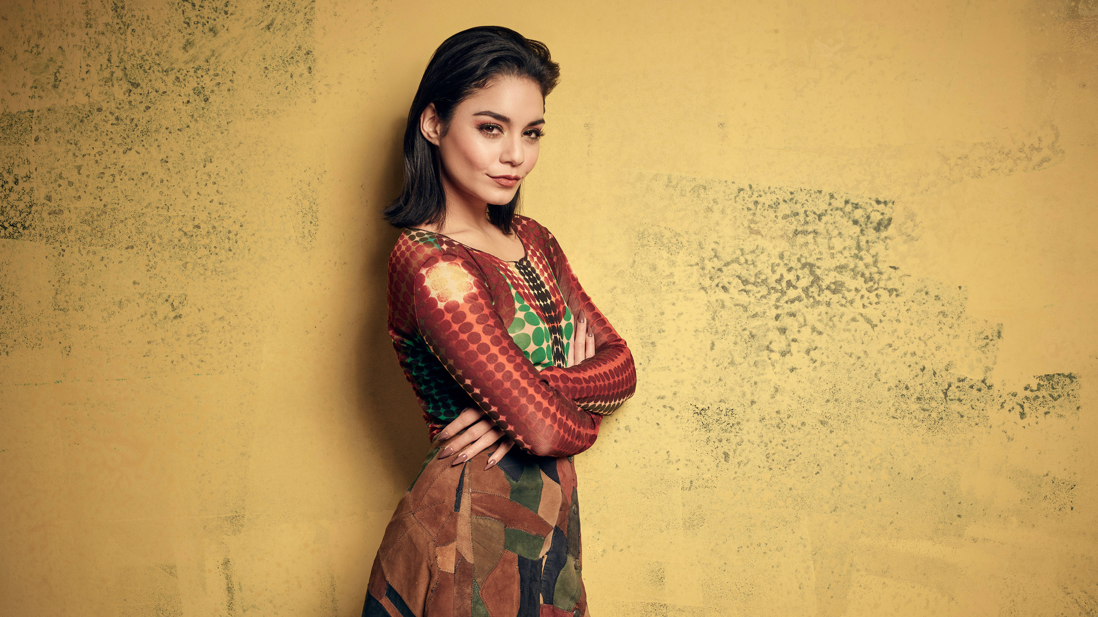 Vanessa Hudgens 桌布 壁紙 — 一位身穿彩色圖案連衣裙的女性自信地交叉雙臂，靠著紋理黃色牆壁站立。