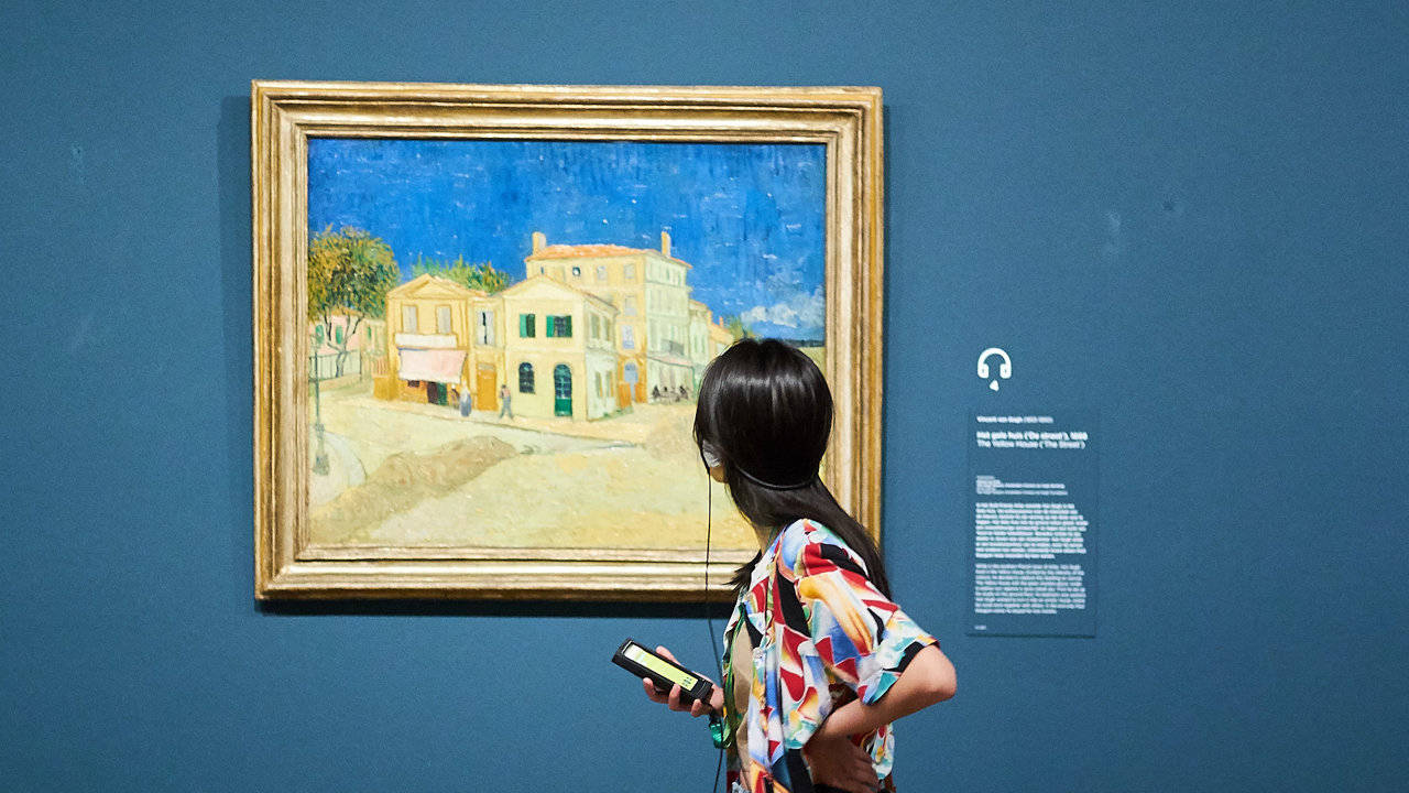 Museum 桌布 壁紙 — 一位身穿彩色襯衫的女子站在博物館畫廊中，面前是一幅梵谷畫作，描繪陽光普照的建築物和鮮豔的藍天。
