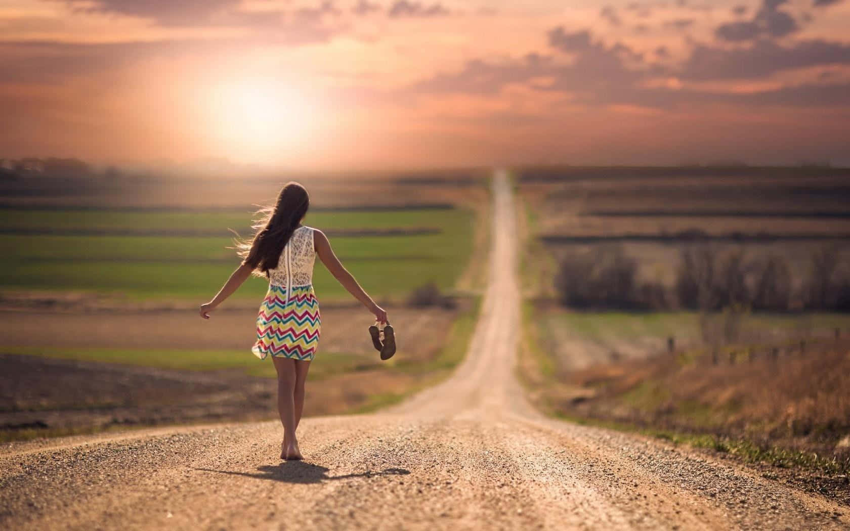 Alone 桌布 壁紙 — 一位身穿彩色連衣裙的女子赤腳沿著一條長長的泥路，走向開闊田野上閃耀的日落。