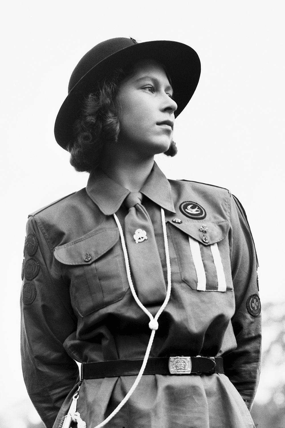 伊莉莎白女王 桌布 壁紙 — 一位身穿復古童軍制服、戴著帽子和繩索領巾的年輕女性，在明亮的天空下向上凝視。