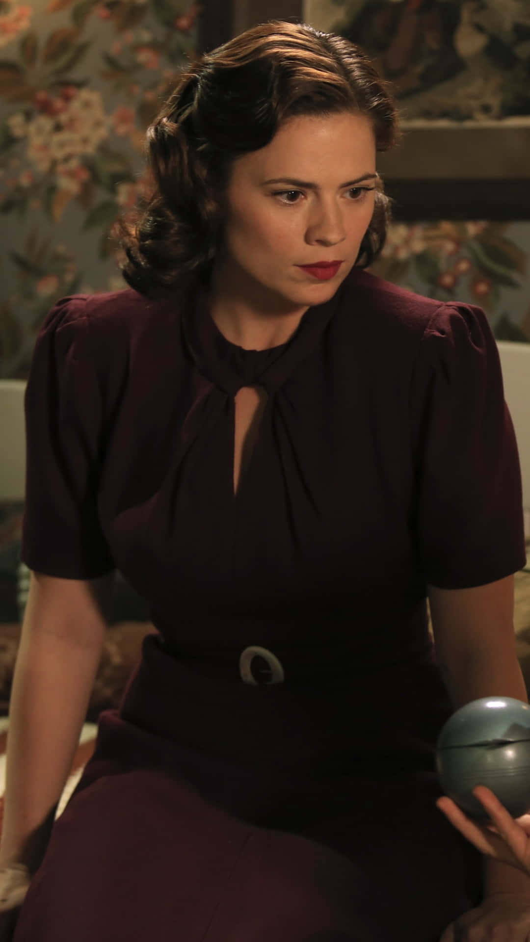 Peggy Carter 桌布 壁紙 — 一位身穿復古酒紅色連衣裙、領口有鑰匙孔設計的女性，坐在花卉壁紙背景前，手持一個金屬球體。