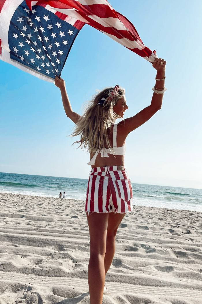 fourth of july 桌布 壁紙 — 一位身穿愛國條紋短褲的女性在陽光明媚的海灘上高舉美國國旗，海面波濤洶湧，天空湛藍清澈。