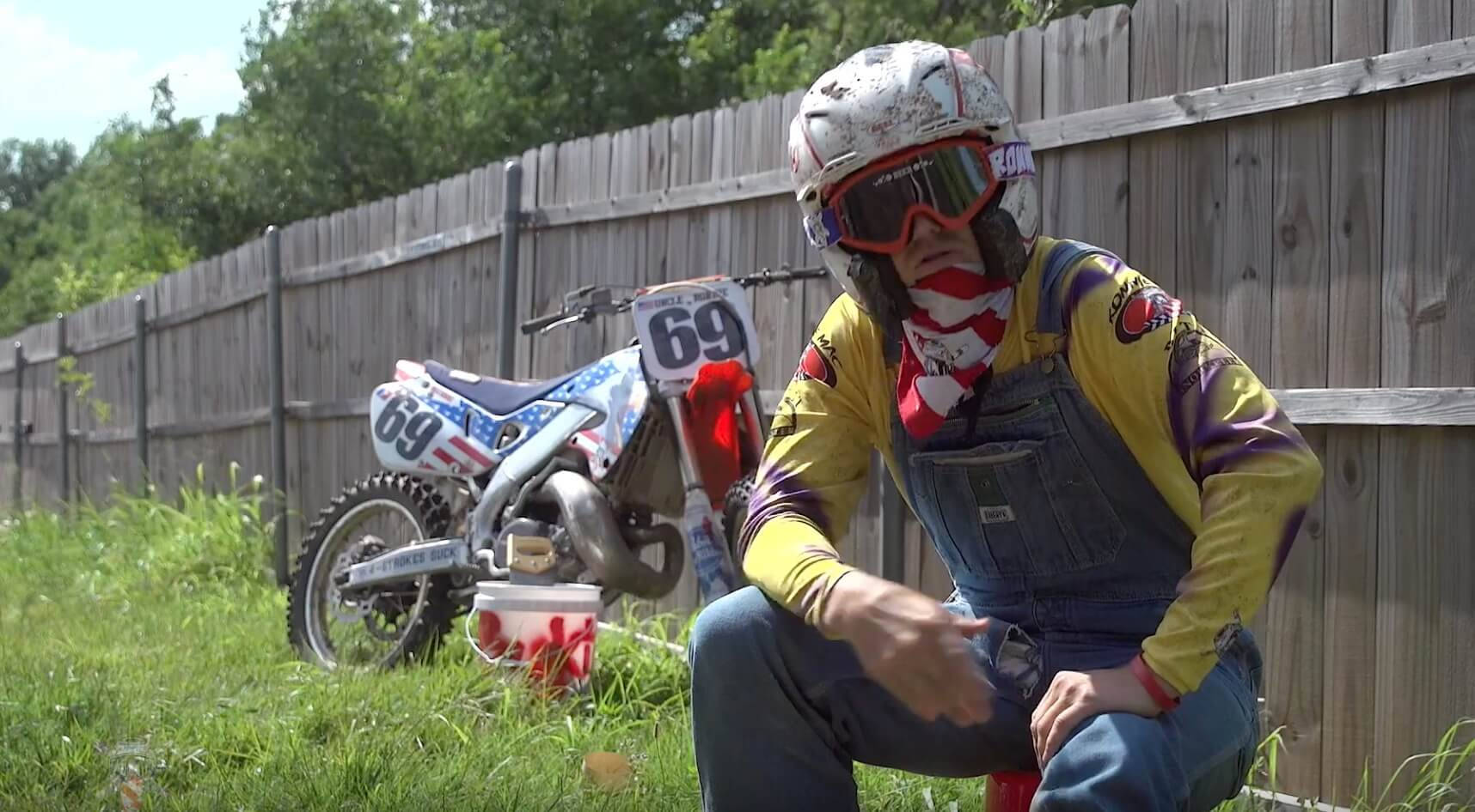 Ronnie Mac 桌布 壁紙 — 一位身穿愛國裝備的越野摩托車手，在陽光明媚的日子裡蹲在他那輛69號摩托車旁，靠近木柵欄。