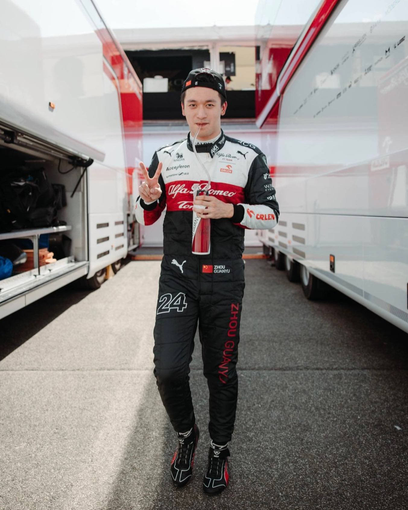 F1 桌布 壁紙 — 一位身穿愛快羅密歐賽車裝備的F1車手微笑著，在兩輛車隊卡車之間拿著一瓶香檳。