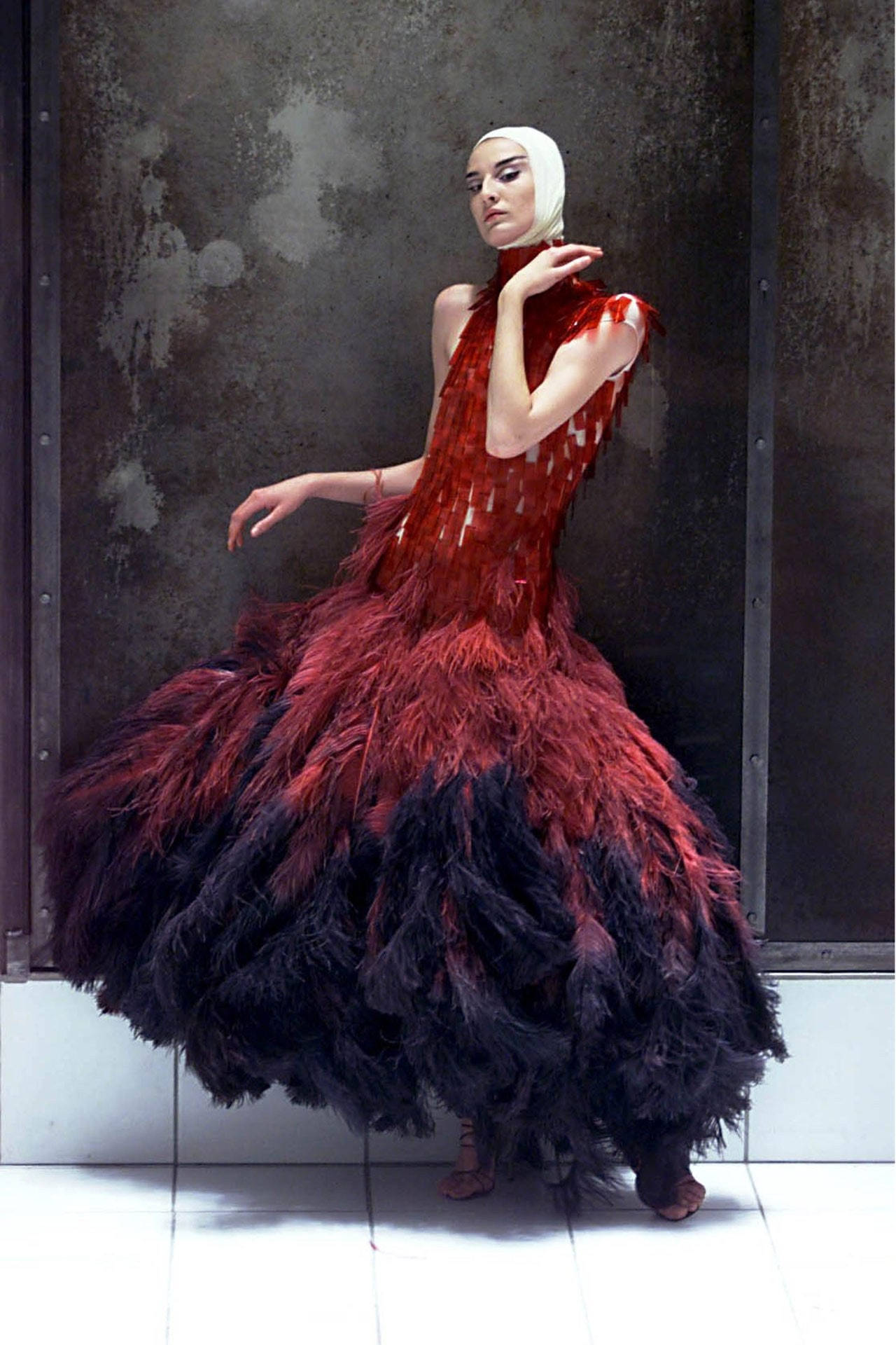 Alexander Mcqueen 桌布 壁紙 — 一位身穿戲劇性紅黑羽毛長裙的女性在風化的工業牆前擺姿勢。