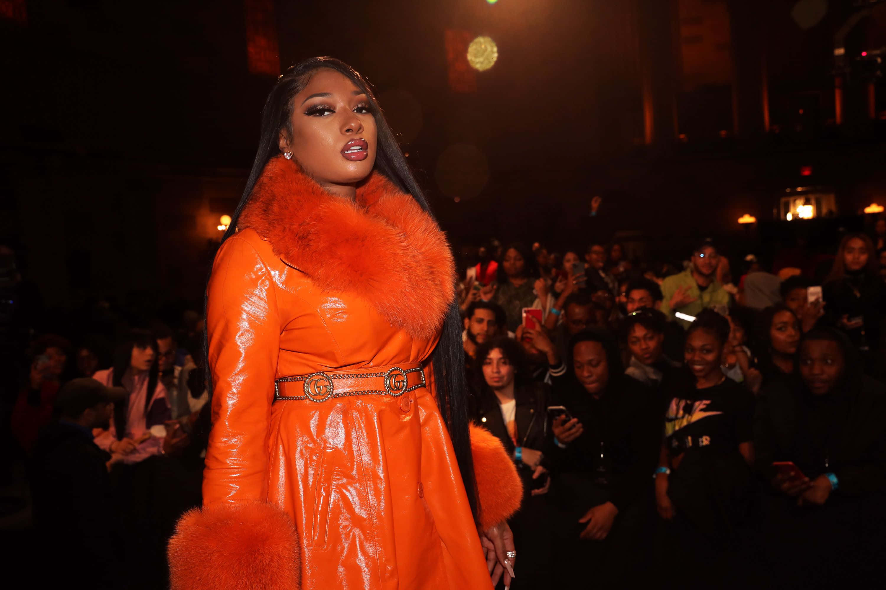Megan Thee Stallion 桌布 壁紙 — 一位身穿搶眼橙色毛邊外套的女性自信地站在燈光昏暗的舞台上，面對觀眾。