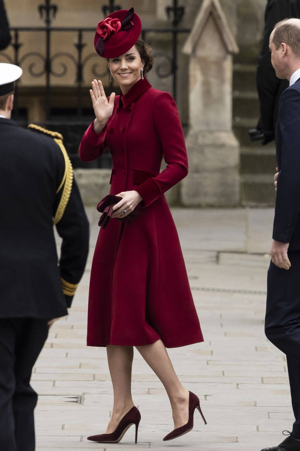 Kate Middleton 桌布 壁紙 — 一位身穿搶眼紅色外套和相配帽子的女士，在石徑上行走時向旁邊的制服人員揮手。