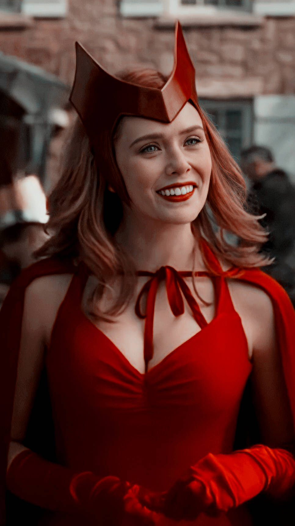 Wanda 桌布 壁紙 — 一位身穿搶眼紅色服裝、頭戴尖冠的女子，在模糊的磚牆建築背景下燦爛微笑。