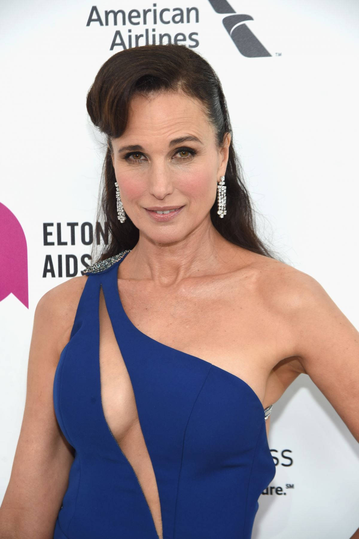 andie macdowell 桌布 壁紙 — 一位身穿搶眼藍色單肩禮服的女士在慈善活動中自信地擺姿勢，身後有贊助商標誌。
