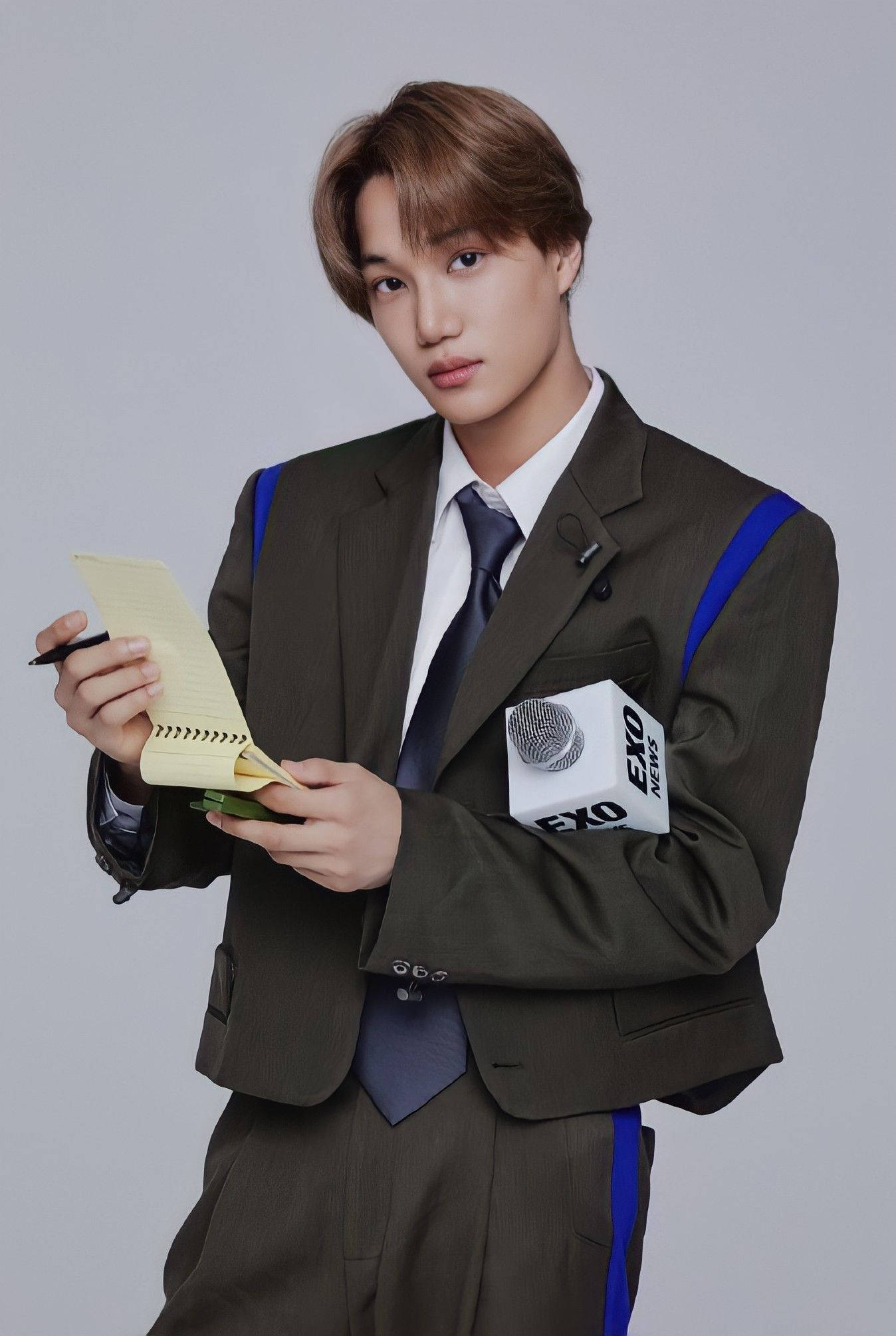 EXO 桌布 壁紙 — 一位身穿時尚棕色西裝、帶有藍色點綴的年輕男子，手持筆記本和筆，直視鏡頭，背景為純灰色。