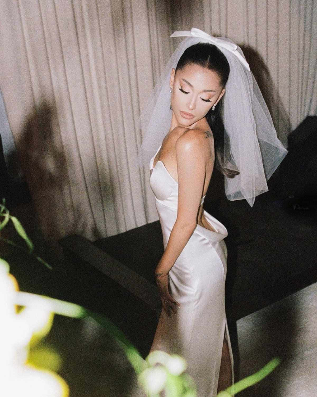 一位身穿白色緞面禮服和頭紗的新娘，在柔和的燈光和模糊的綠色植物中室內擺姿勢。 — 亞莉安娜·格蘭德 GIF 壁紙
