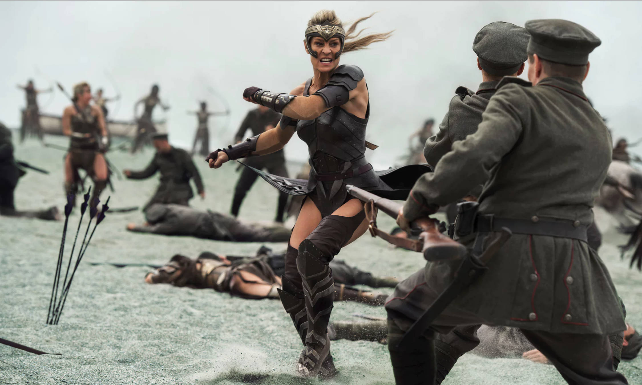 Wonder Woman 桌布 壁紙 — 一位身穿暗色鎧甲的兇猛女戰士在混亂的戰場上與士兵作戰，灰色地面上散落著倒下的戰士和箭矢。
