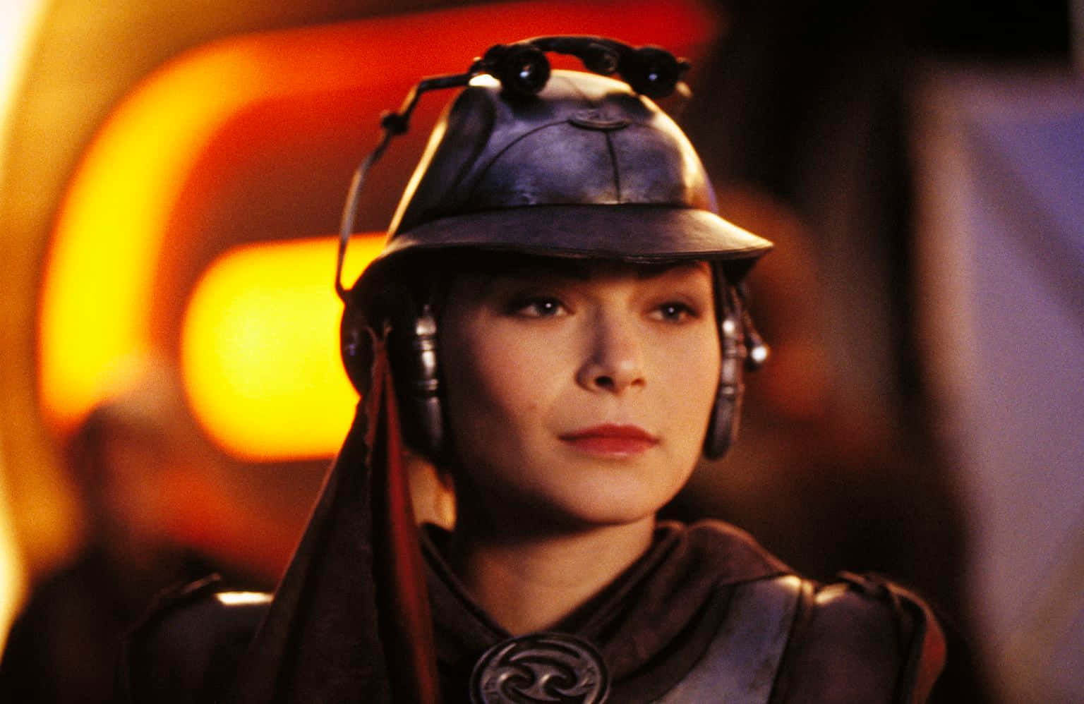 Zam Wesell 桌布 壁紙 — 一位身穿未來風格深色盔甲和頭盔的女性，在溫暖的橙色光芒下凝視前方。