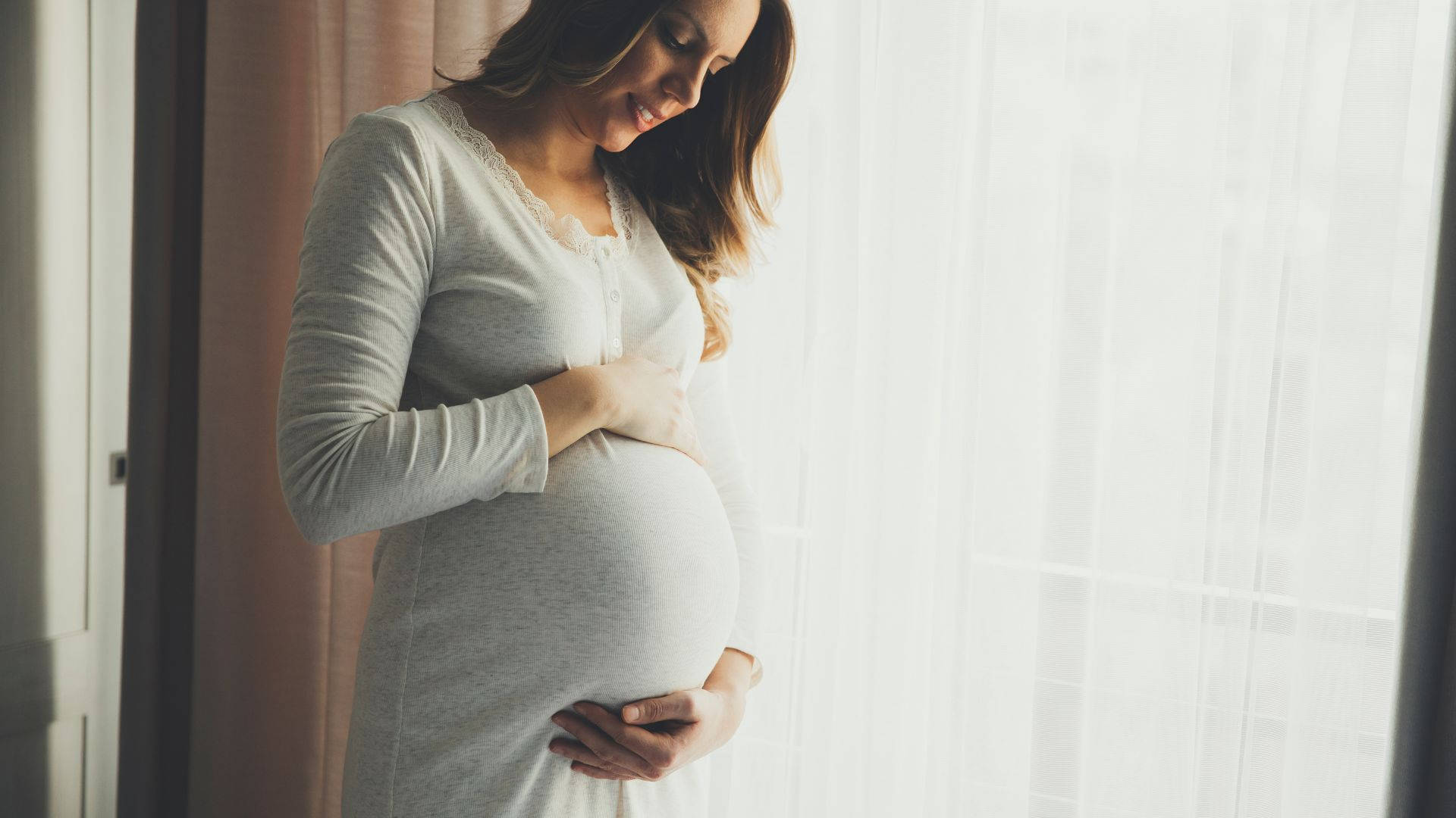 Woman 桌布 壁紙 — 一位身穿柔和灰色連衣裙的孕婦，站在明亮窗邊，輕柔地抱著肚子，窗簾輕薄透光。