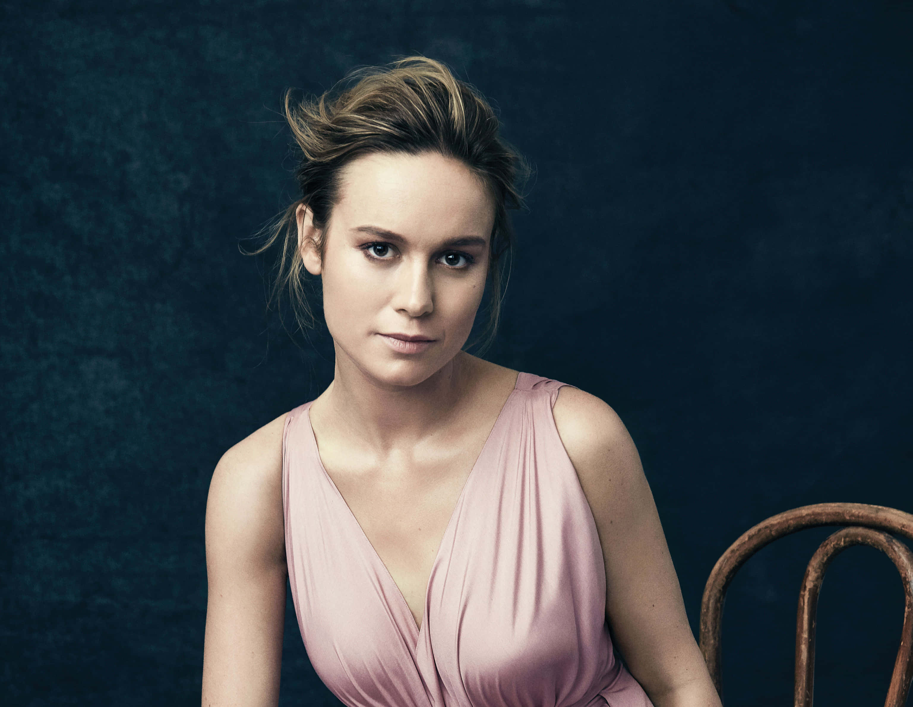 brie larson 桌布 壁紙 — 一位身穿柔和粉紅色連衣裙的女性優雅地坐在質感深藍色背景前，旁邊有一張質樸的木椅。