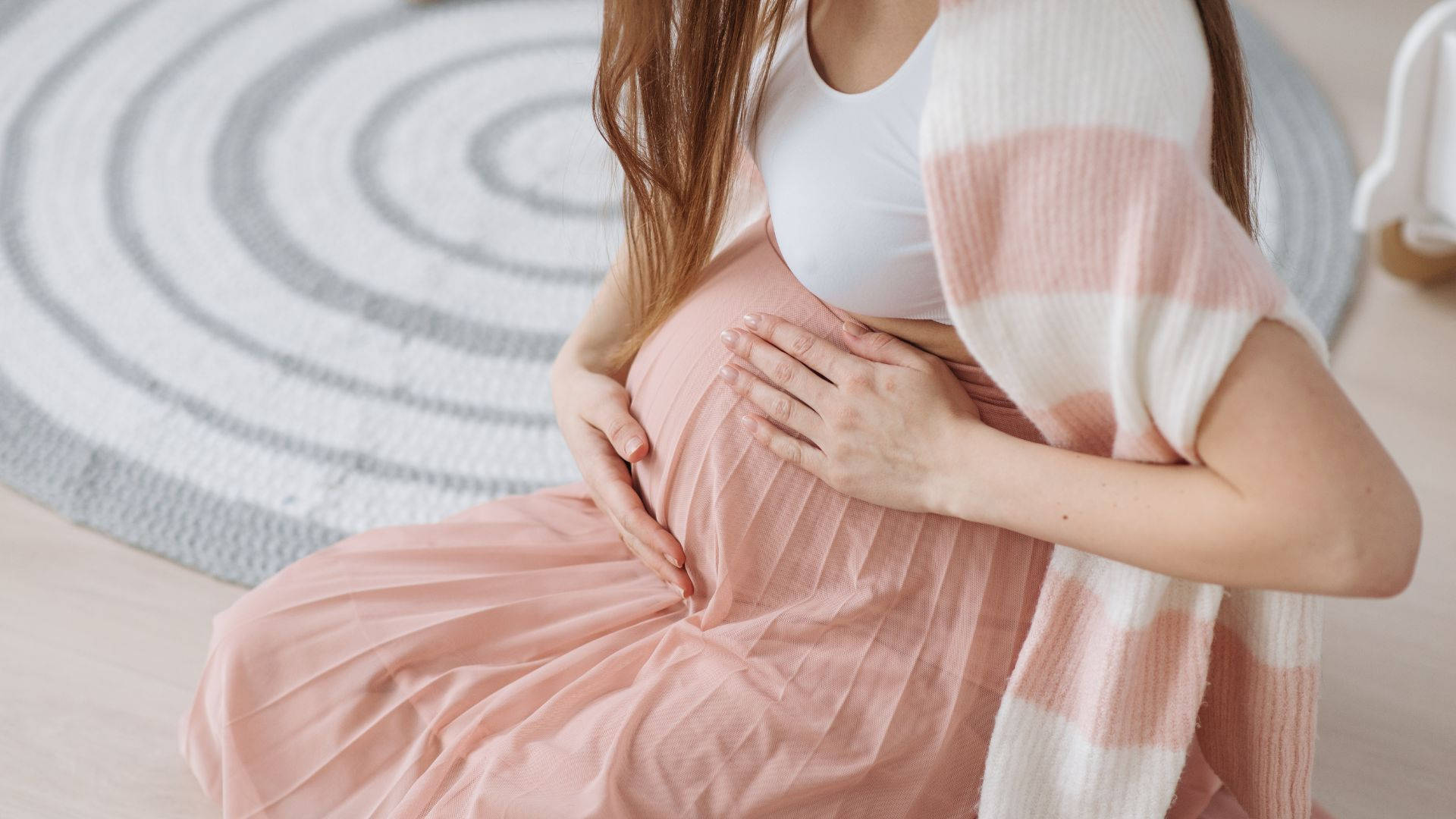 Woman 桌布 壁紙 — 一位身穿柔和粉紅色連衣裙的孕婦，在室內螺旋形地毯上輕輕地抱著肚子。