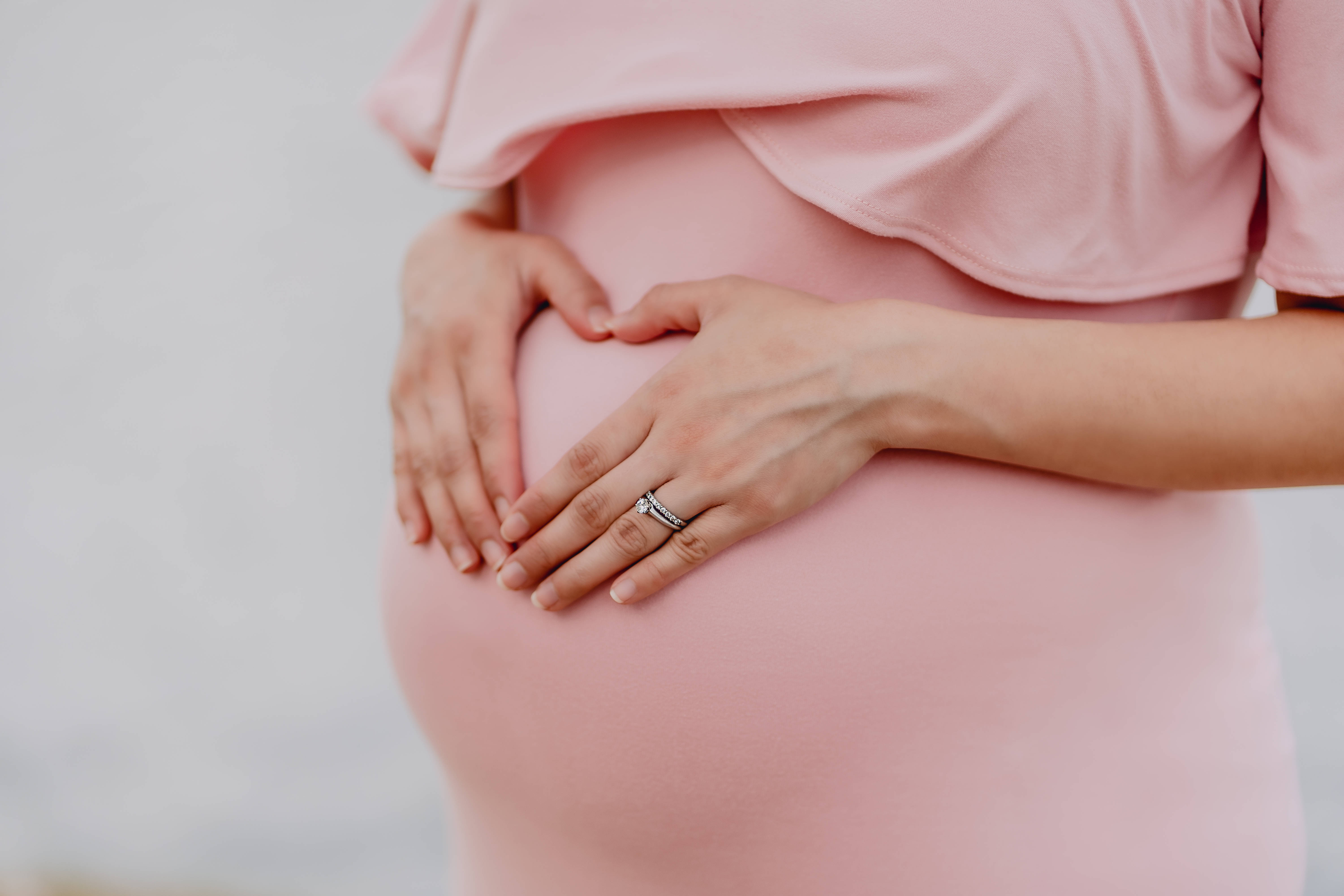 Pregnant 桌布 壁紙 — 一位身穿柔和粉紅色連衣裙的孕婦，雙手輕輕捧著腹部，形成心形。