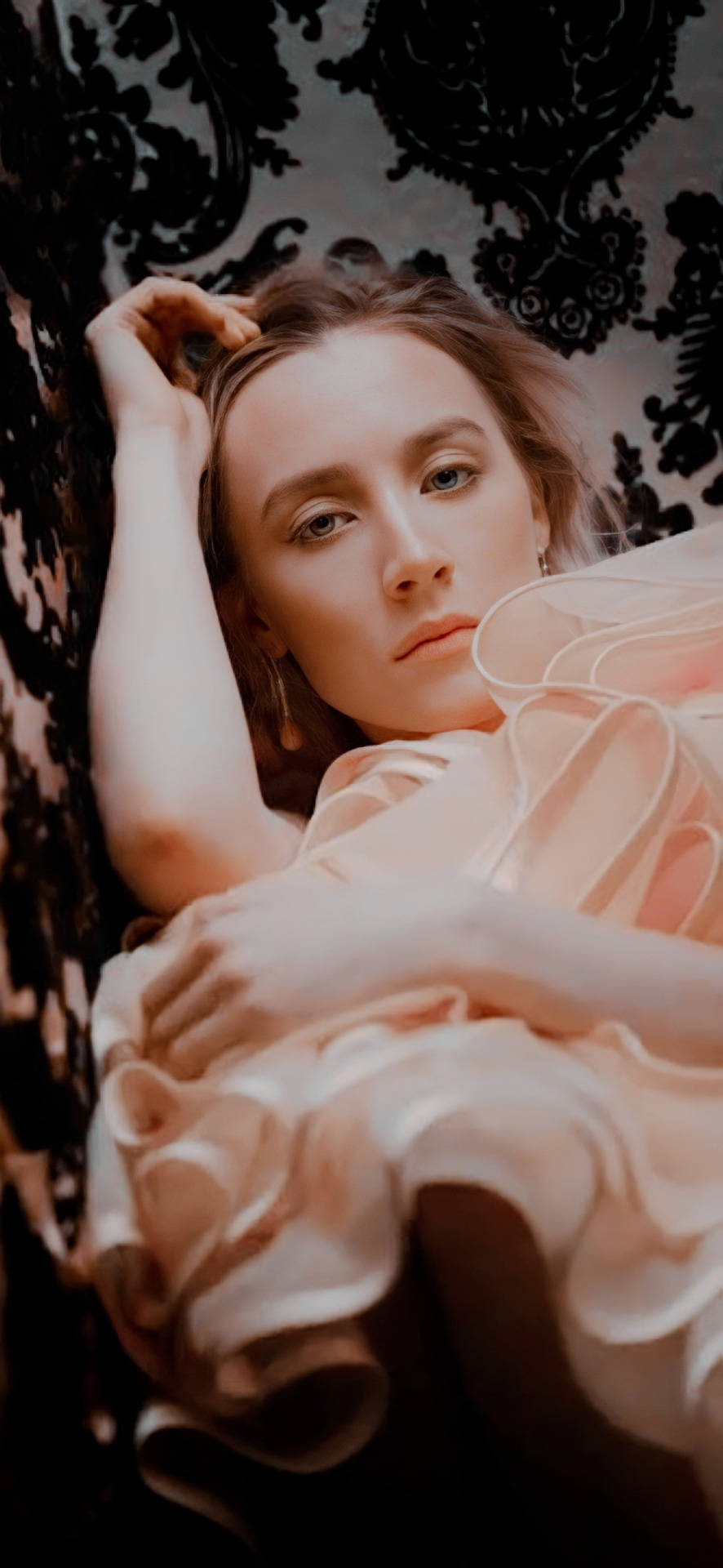 Saoirse Ronan 桌布 壁紙 — 一位身穿柔和蜜桃色長袍的女性，倚靠在黑色花卉壁紙上，眼神若有所思。