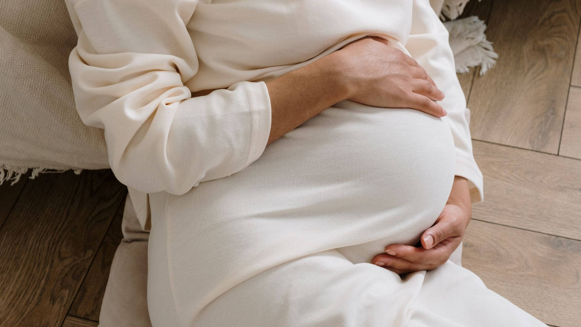 Woman 桌布 壁紙 — 一位身穿柔軟白色連衣裙的孕婦，坐在舒適的質感沙發上，輕輕地抱著她的肚子。