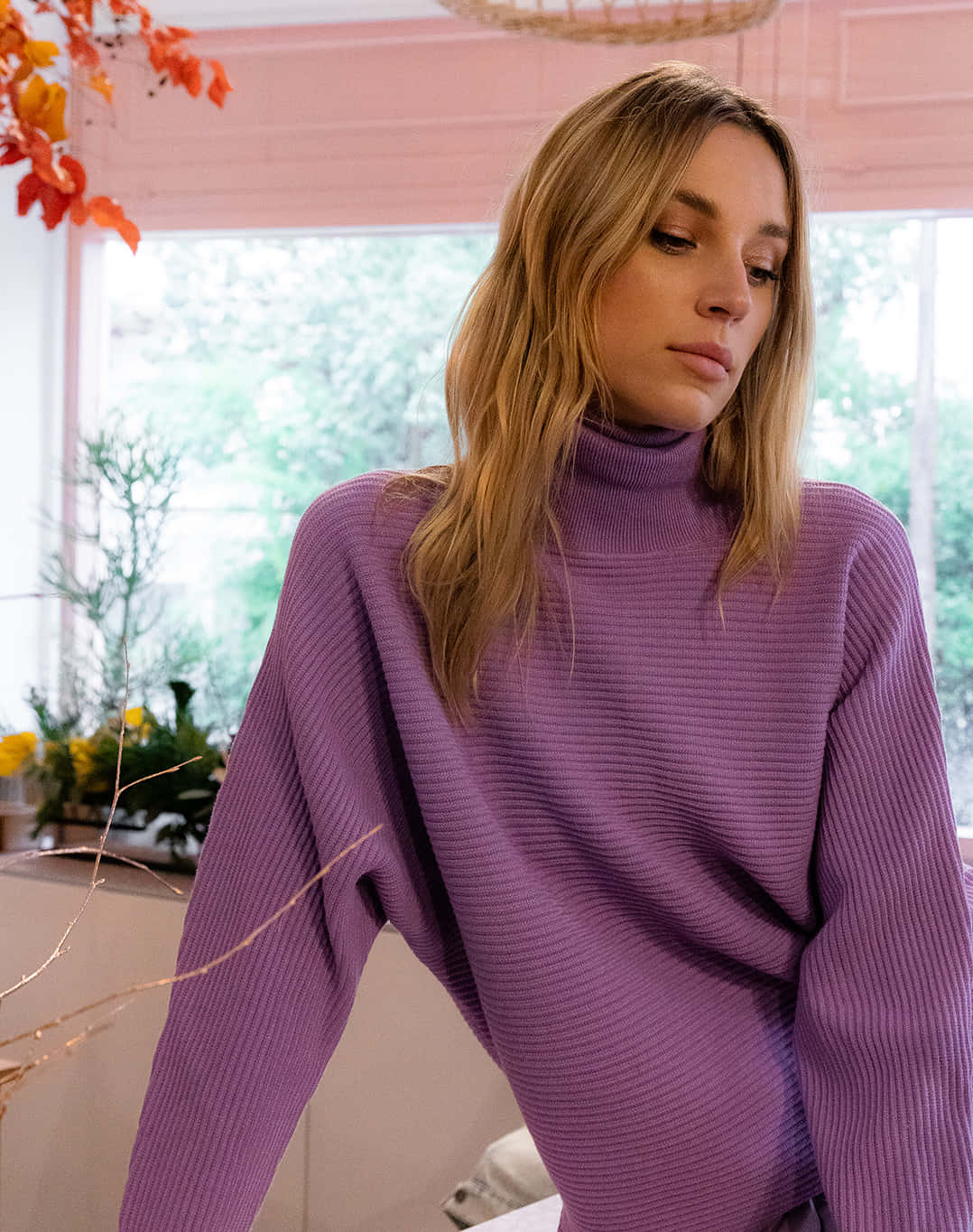 Purple Turtle-neck Sweater 桌布 壁紙 — 一位身穿柔軟紫色羅紋高領毛衣的女性站在室內靠近窗邊，窗外有秋葉和綠植。