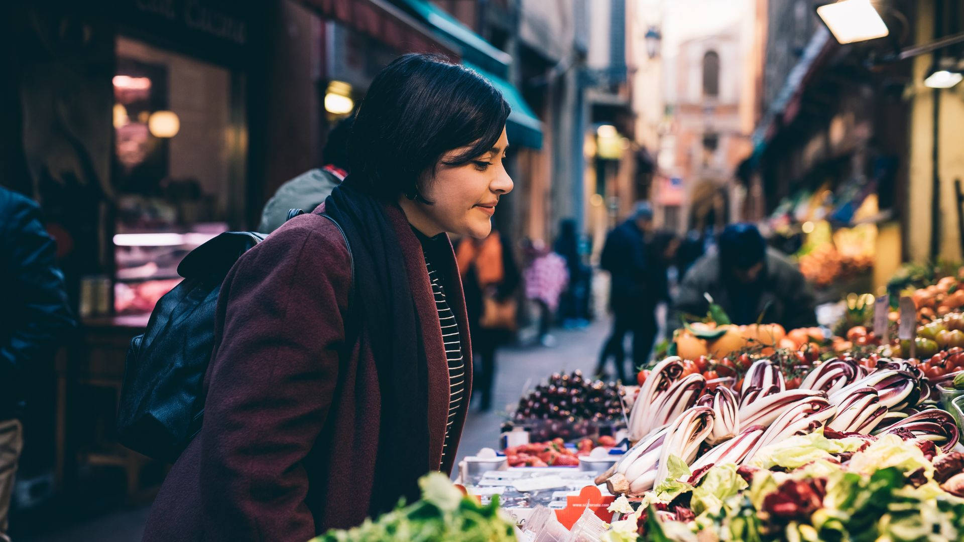 Fruits 桌布 壁紙 — 一位身穿栗色外套的女性在熱鬧的歐洲街市瀏覽色彩繽紛的新鮮農產品時微笑。