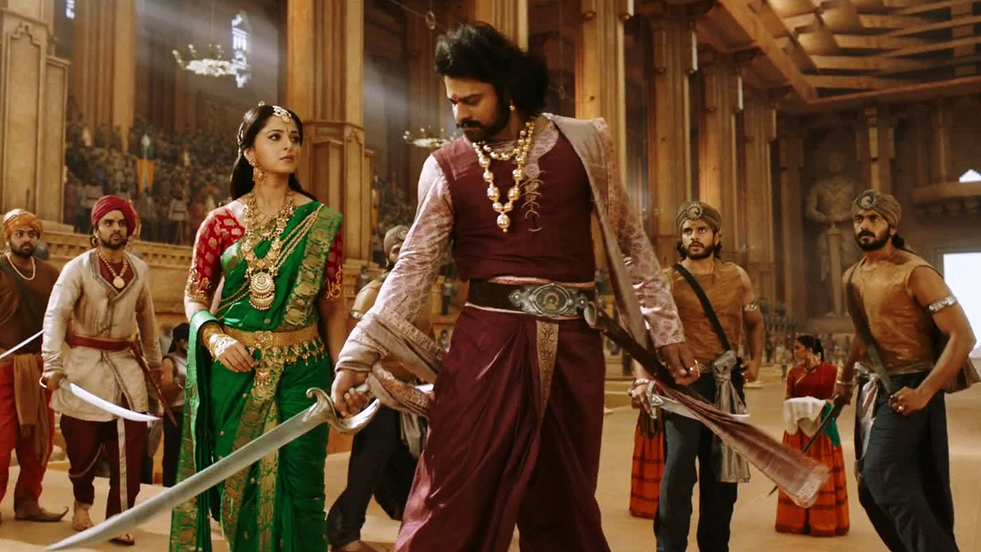 Prabhas HD 桌布 壁紙 — 一位身穿栗色多蒂和金色珠寶的威嚴戰士手持劍向前邁進，兩側是一位穿著綠色紗麗的女子和武裝隨從，在華麗的宮殿大廳中。