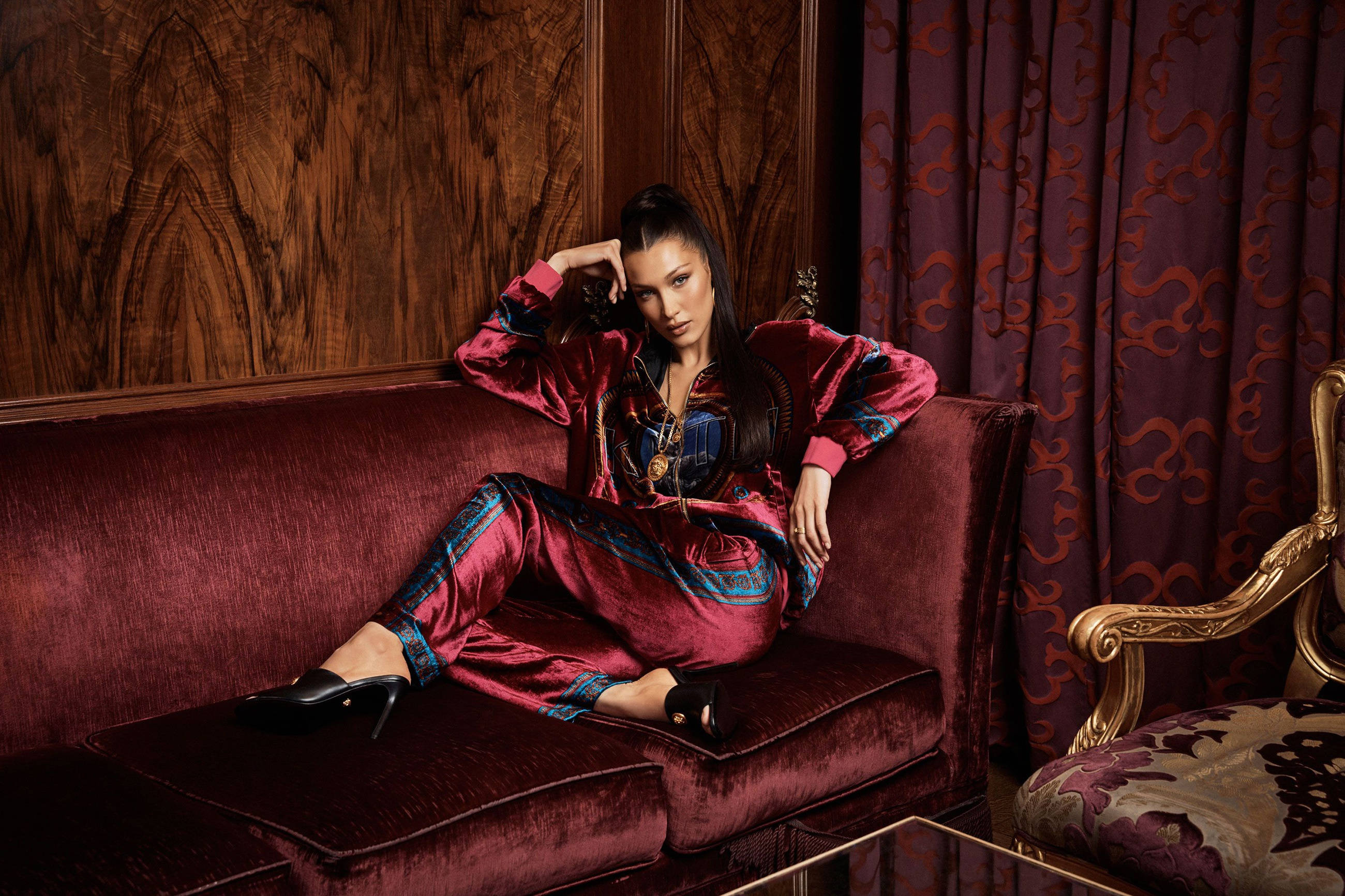 Bella Hadid 桌布 壁紙 — 一位身穿栗色天鵝絨家居服的女士，在一個裝飾華麗、帶有華麗木鑲板和紫色窗簾的房間裡，斜倚在相配的沙發上。