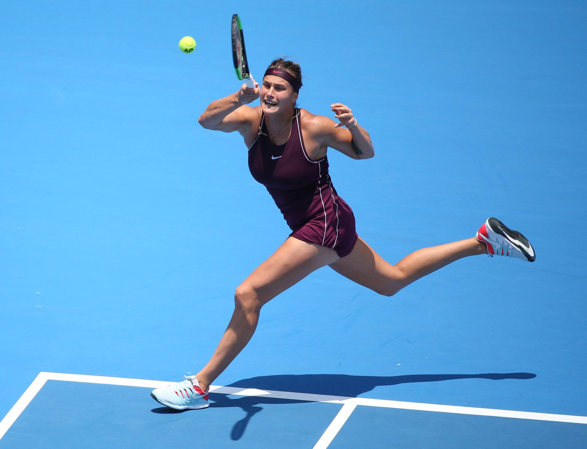 Aryna Sabalenka 桌布 壁紙 — 一位身穿栗色裝備的女網球運動員在晴朗的日光下，在鮮豔的藍色球場上動態地弓步擊打一個亮黃色的球。