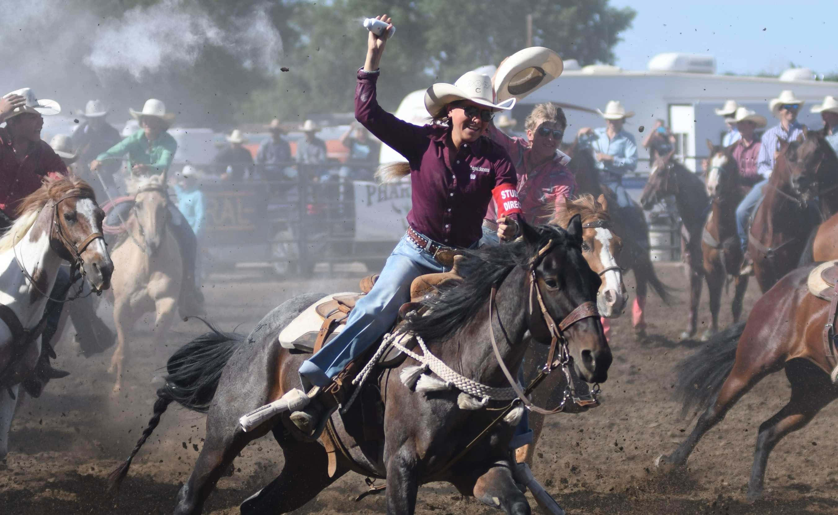 Rodeo 桌布 壁紙 — 一位身穿栗色襯衫、頭戴白帽的牛仔，在塵土飛揚的牛仔競技中，在一匹騰躍的黑馬上勝利地舉起手臂。