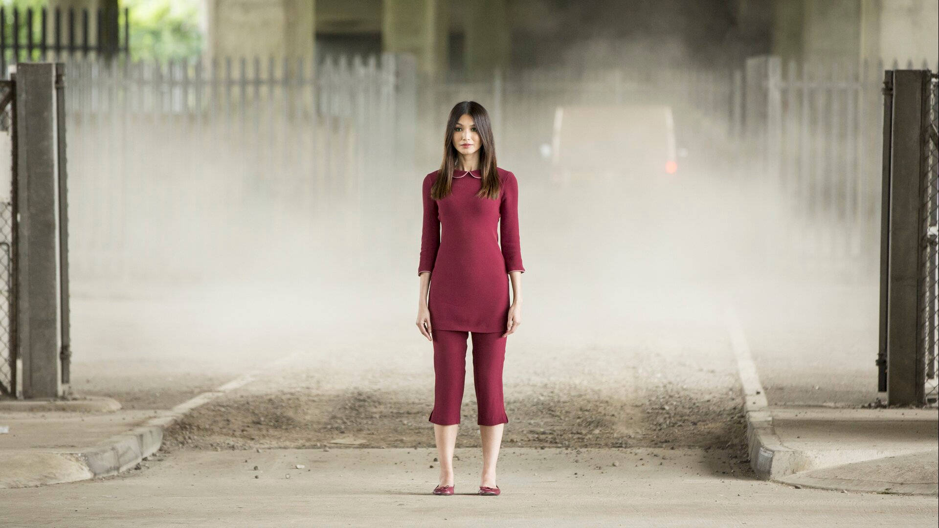 Gemma Chan 桌布 壁紙 — 一位身穿栗色連衣裙的女性獨自站在一個佈滿灰塵的城市小巷中，周圍是鐵絲網圍欄和混凝土結構。