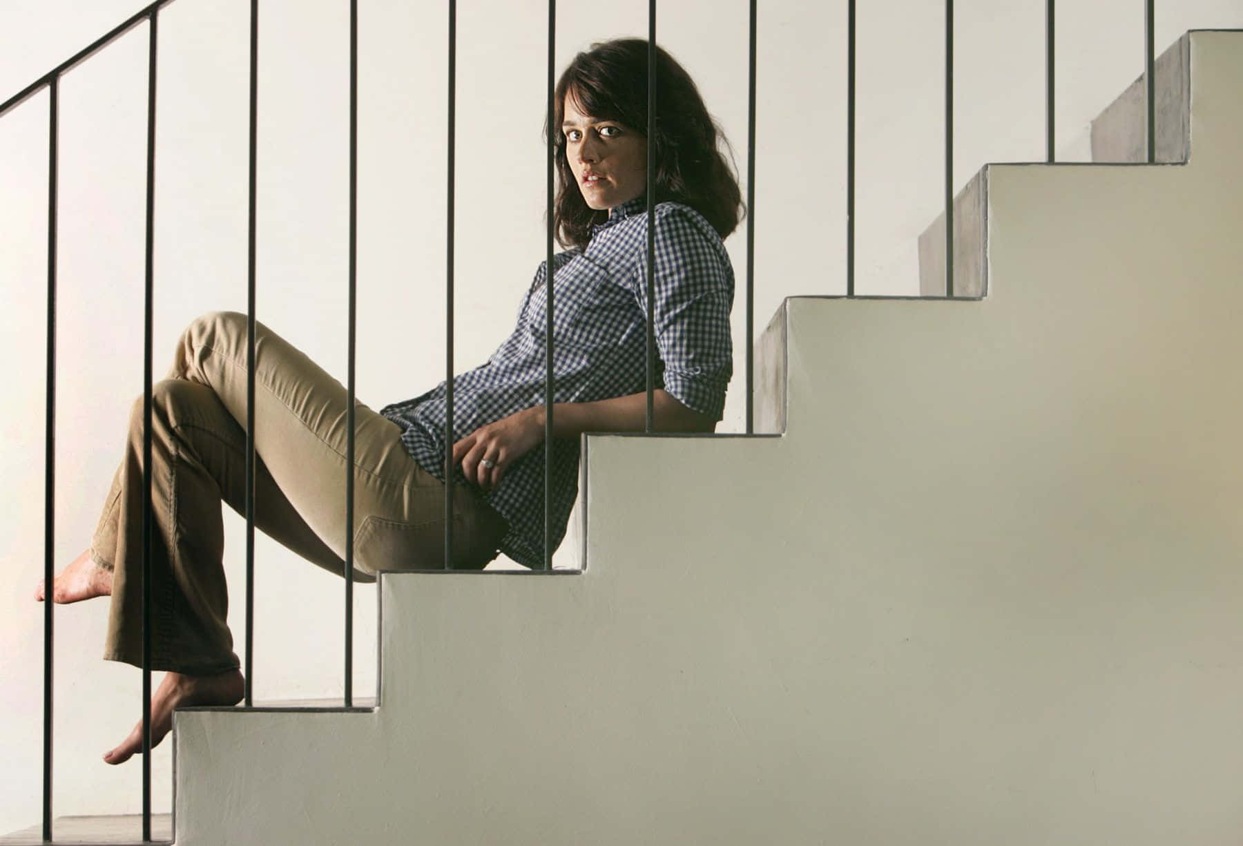 Robin Tunney 桌布 壁紙 — 一位身穿格子襯衫的女人赤腳坐在由黑色金屬欄杆框住的簡約混凝土樓梯上。