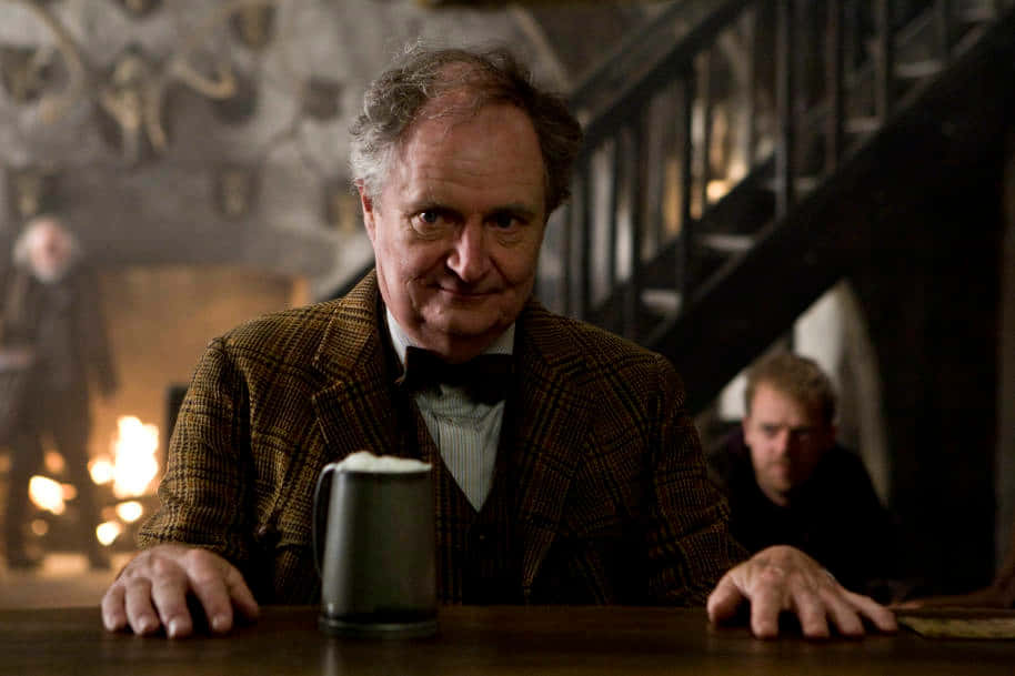 The Hogwarts Professors 桌布 壁紙 — 一位身穿格紋外套和領結的年長男士，靠著木製吧檯，面前放著一杯啤酒，對著鏡頭微笑。