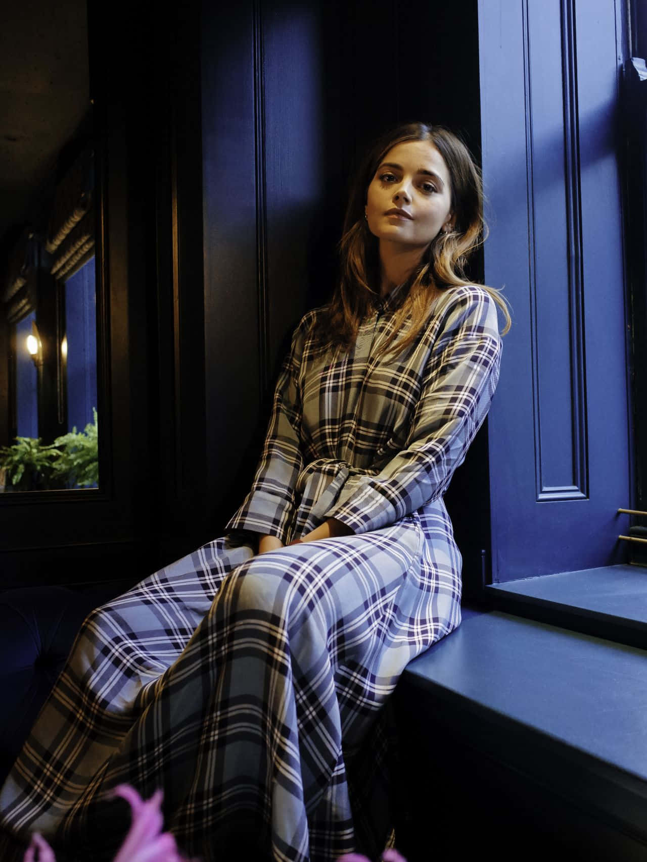 Jenna Coleman 桌布 壁紙 — 一位身穿格紋裙的女子沉思地坐在藍色窗台上，沐浴在柔和的晚光中。