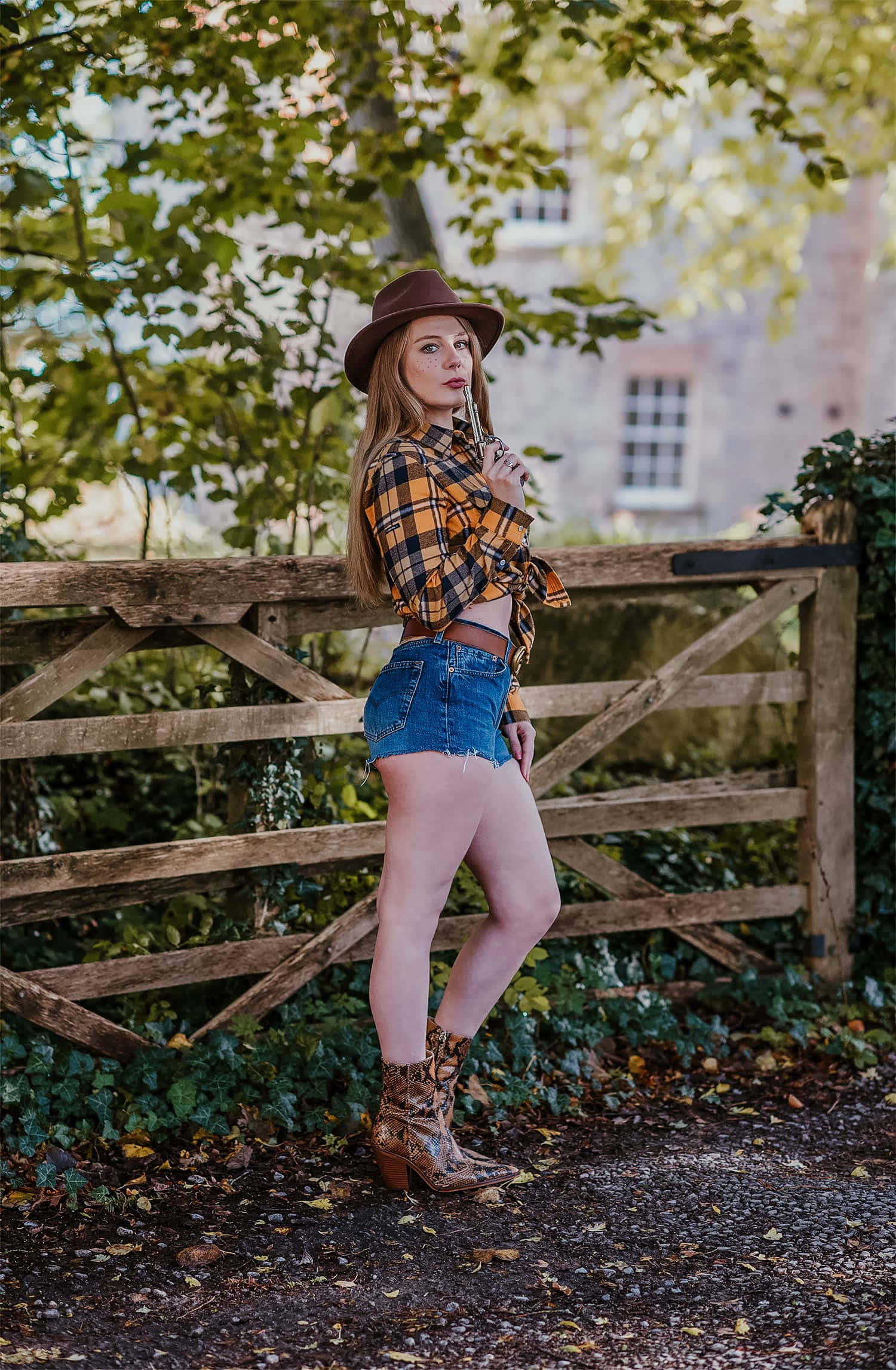 Country Girl 桌布 壁紙 — 一位身穿格紋襯衫、牛仔短褲和牛仔靴的女性，在枝葉茂密的樹下自信地靠在木柵欄旁擺姿勢。