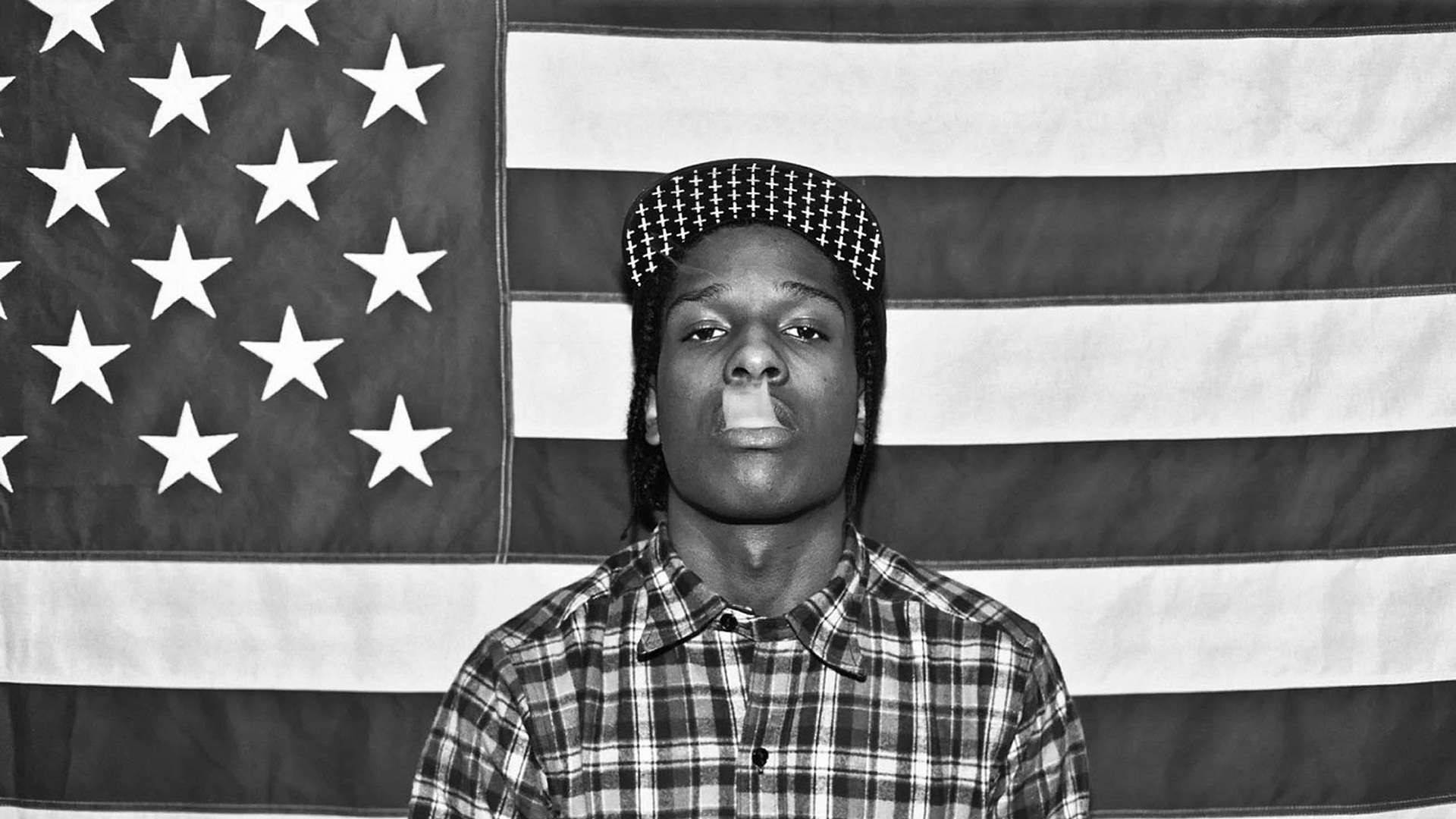 Rap 桌布 壁紙 — 一位身穿格紋襯衫和格紋帽的年輕男子自信地站在黑白美國國旗前。