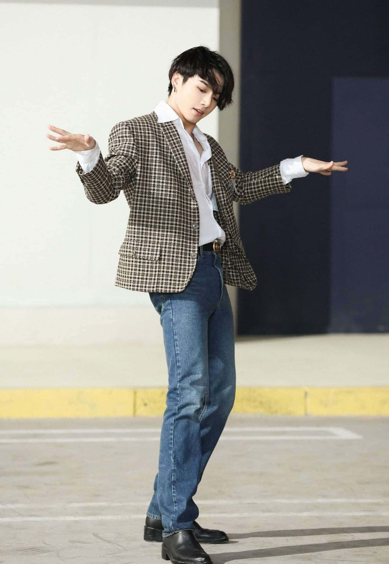 Jungkook 桌布 壁紙 — 一位身穿格紋西裝外套和牛仔褲的年輕男子，在極簡城市背景下伸展雙臂，擺出充滿動感的姿勢。