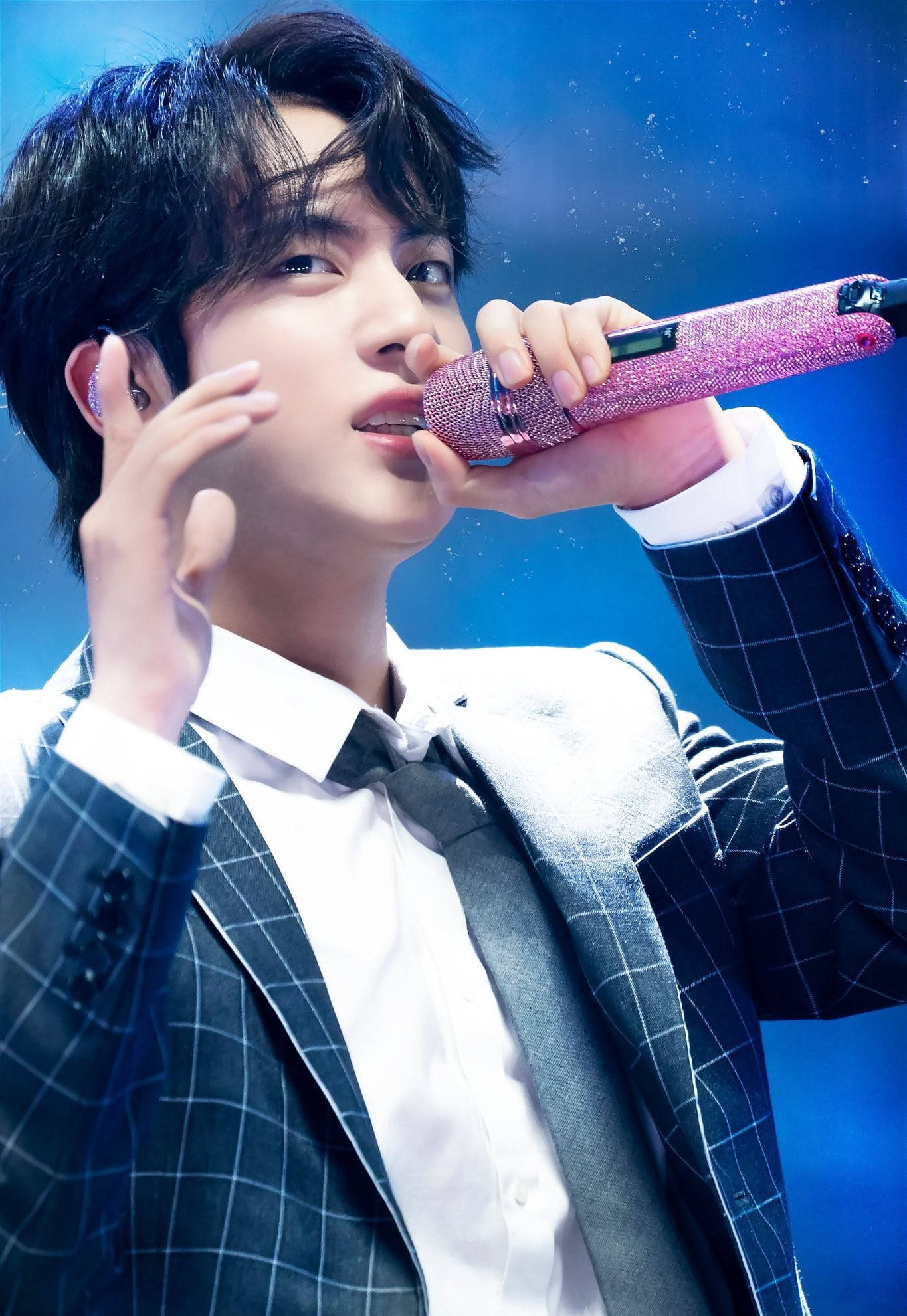 kim seok jin 桌布 壁紙 — 一位身穿格紋西裝的男歌手，在充滿活力的藍色舞台燈光下，熱情地對著閃閃發光的粉紅色麥克風演唱。