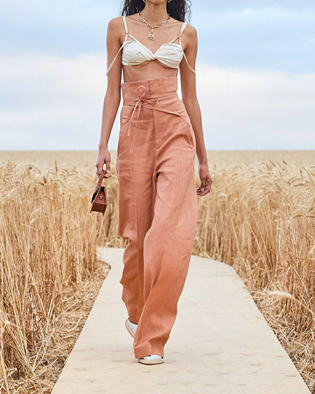 Wheat Field 桌布 壁紙 — 一位身穿桃色亞麻褲和白色上衣的女性，自信地走在柔和的藍天下，穿過金色麥田的木質小徑上。