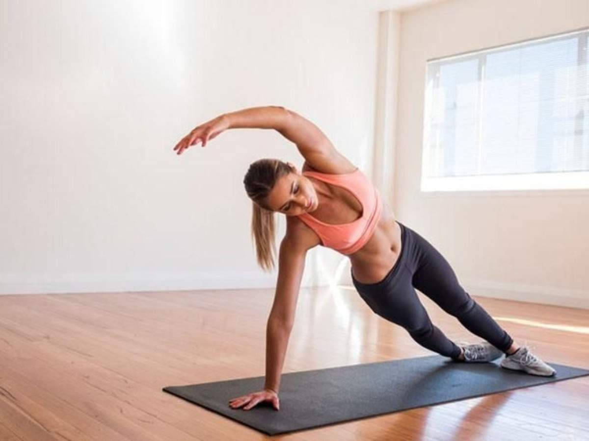 Exercise 桌布 壁紙 — 一位身穿桃色運動胸罩和黑色緊身褲的女性，在鋪有木地板和大型窗戶的明亮簡約房間內的黑色瑜伽墊上進行側平板支撐伸展。