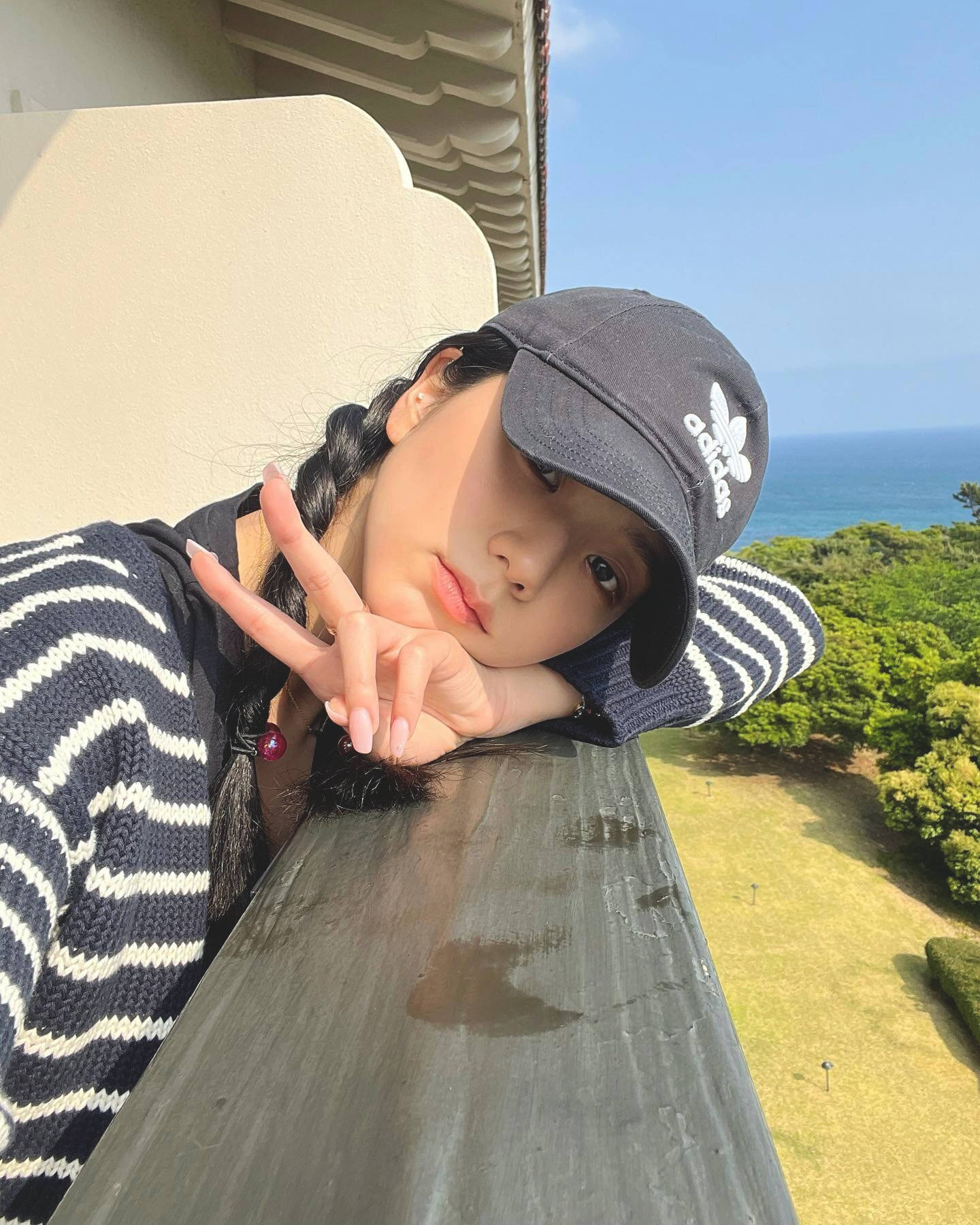 jisoo 可愛 桌布 壁紙 — 一位身穿條紋毛衣和帽子的年輕女子靠在陽台欄杆上，在清澈藍天下以海洋和綠色山丘為背景做出和平手勢。