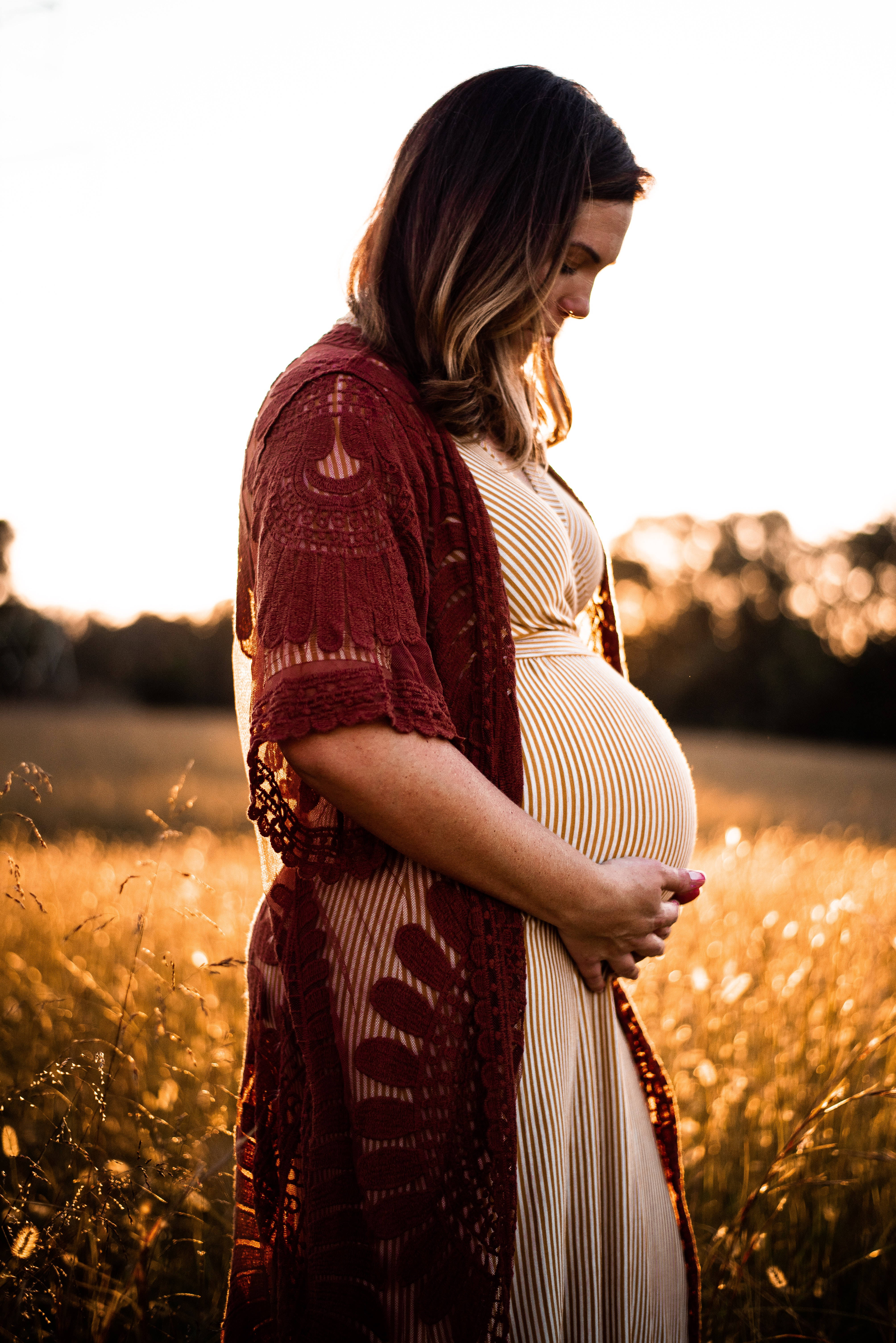 Pregnant 桌布 壁紙 — 一位身穿條紋洋裝和紅色蕾絲開襟衫的孕婦，在夕陽下的金色田野中輕輕地捧著她的肚子。