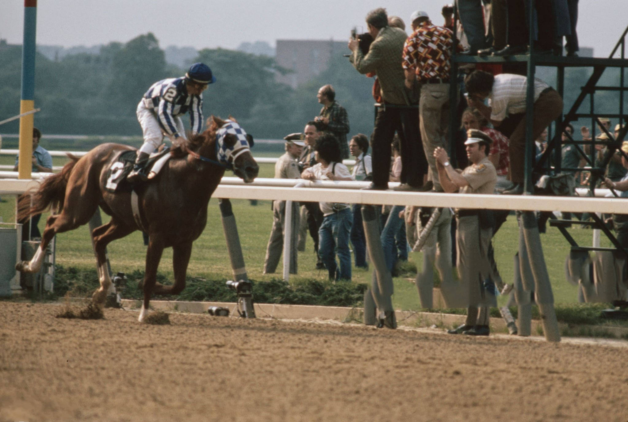 Secretariat 桌布 壁紙 — 一位身穿條紋絲綢的騎師騎著一匹棕色的二號賽馬，從泥地賽道的閘門衝出，觀眾在旁觀看。