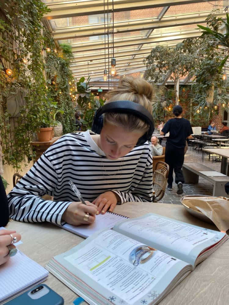 書桌 桌布 壁紙 — 一位身穿條紋襯衫、戴著耳機的年輕女子，在鬱鬱蔥蔥的室內花園咖啡館的木桌上專心學習。