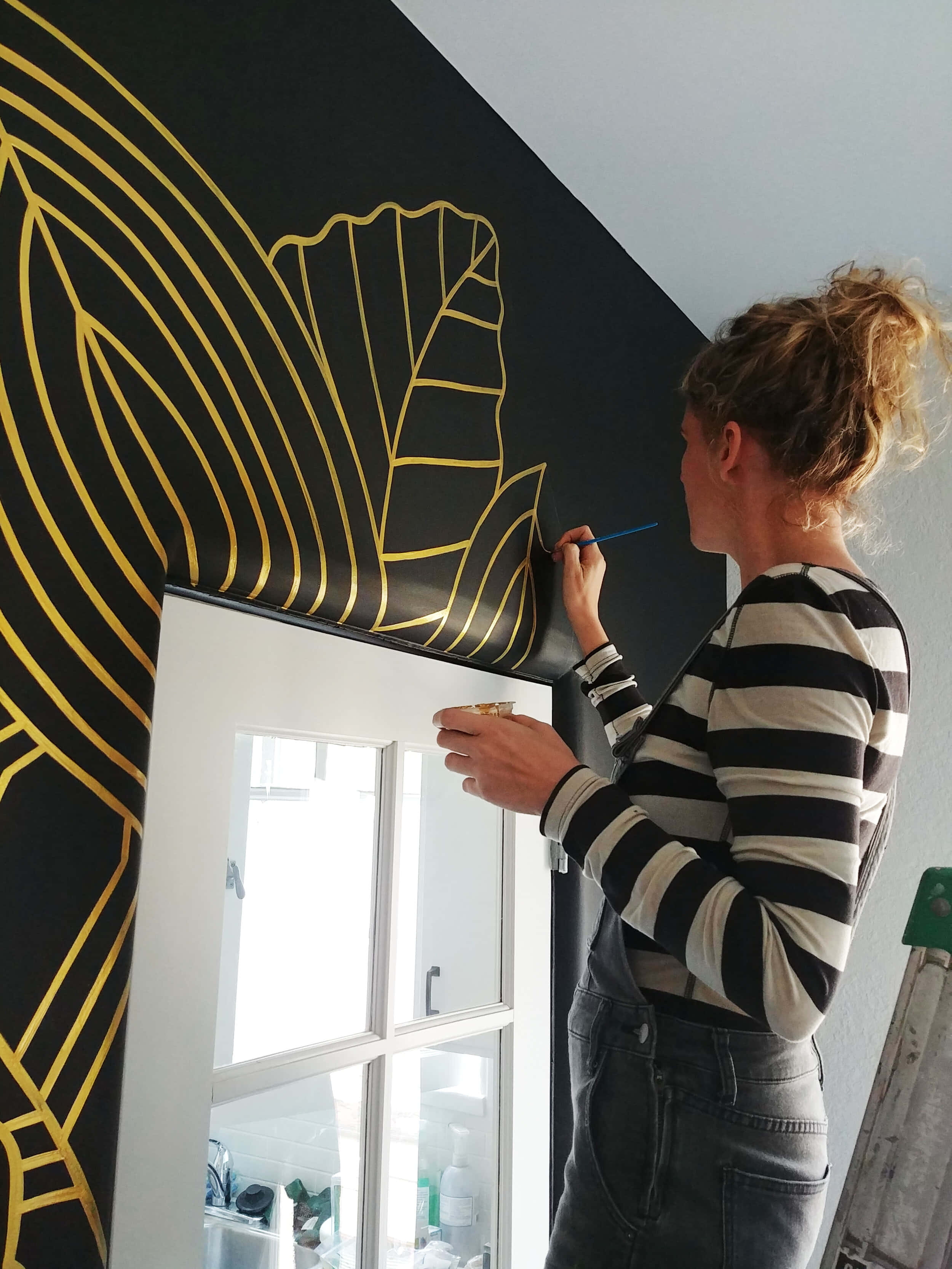 Wall Painting 桌布 壁紙 — 一位身穿條紋襯衫的女性正在白色窗框旁邊的黑色牆壁上仔細繪畫金色葉子圖案。