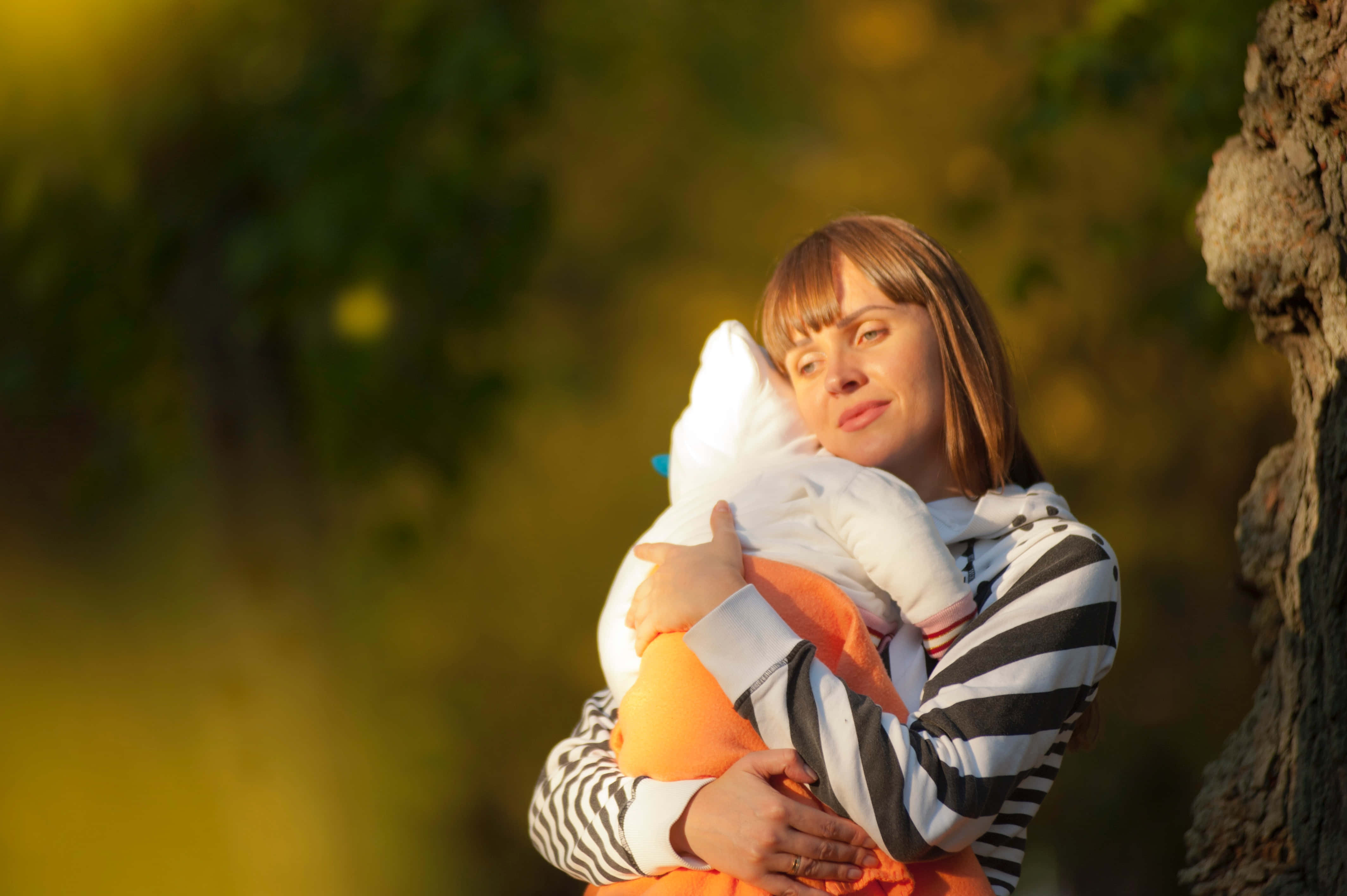 Mom 桌布 壁紙 — 一位身穿條紋連帽衫的女士，在陽光普照、金黃樹葉的樹幹旁，溫柔地抱著一個裹著白布的嬰兒。