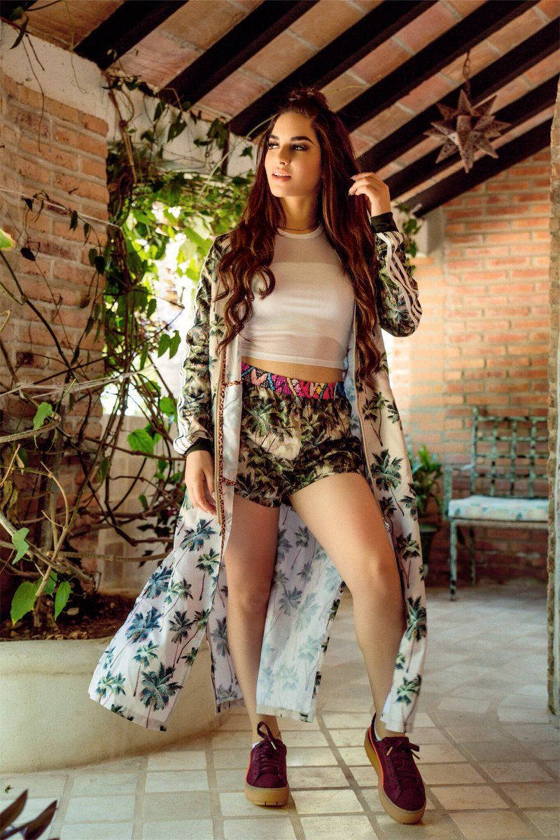 Latina Girl 桌布 壁紙 — 一位身穿棕櫚印花和服和短褲的女性自信地站在一個有懸掛藤蔓和木樑的鄉村磚砌庭院下。